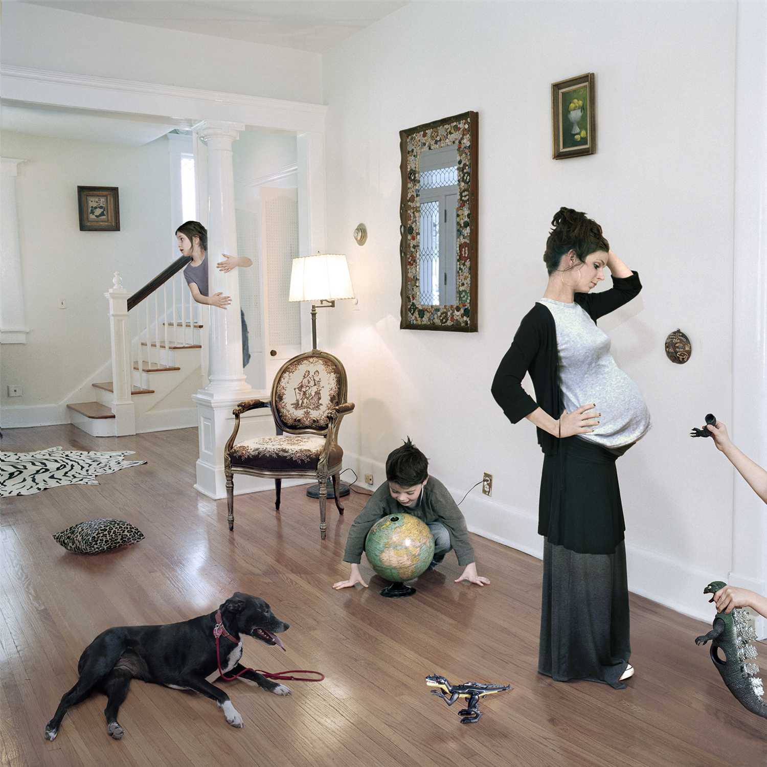 A pregnant woman stands in a bright, traditionally decorated home interior, one hand on her belly and the other to her head in a gesture of bewilderment. Around her, children and a black dog engage in playful chaos—a child crawls with a globe, another leans over a staircase railing, toys and objects scatter across the wooden floor. The composition captures a moment of domestic life filled with simultaneous activity and disorder.