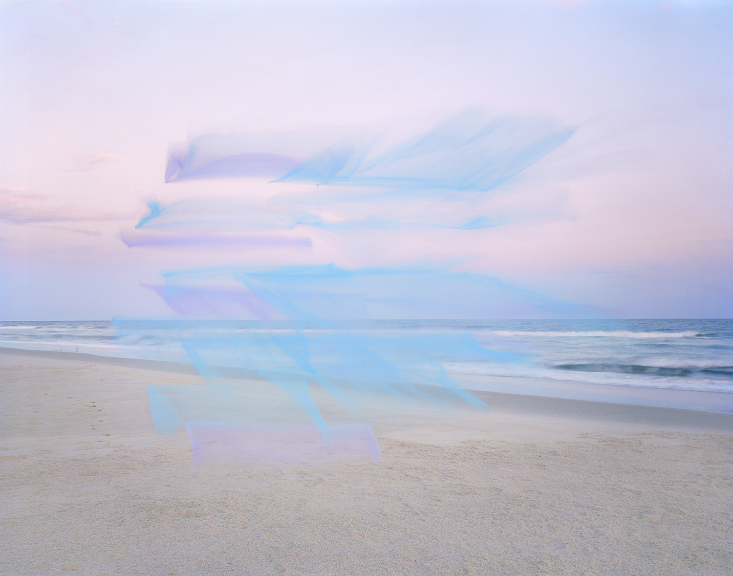 Translucent blue and purple tulle fabric overlays a serene beach landscape at dusk, with soft sand in the foreground and calm ocean waters meeting a pale sky at the horizon.