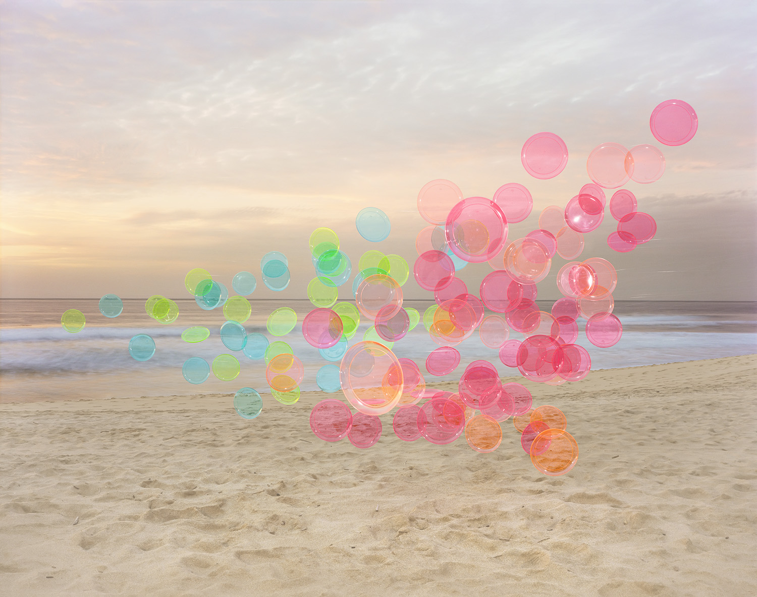 Translucent colored spheres in shades of pink, yellow, blue, and orange arranged in a floating cluster above a sandy beach at sunset, with calm ocean and soft sky visible in the background.