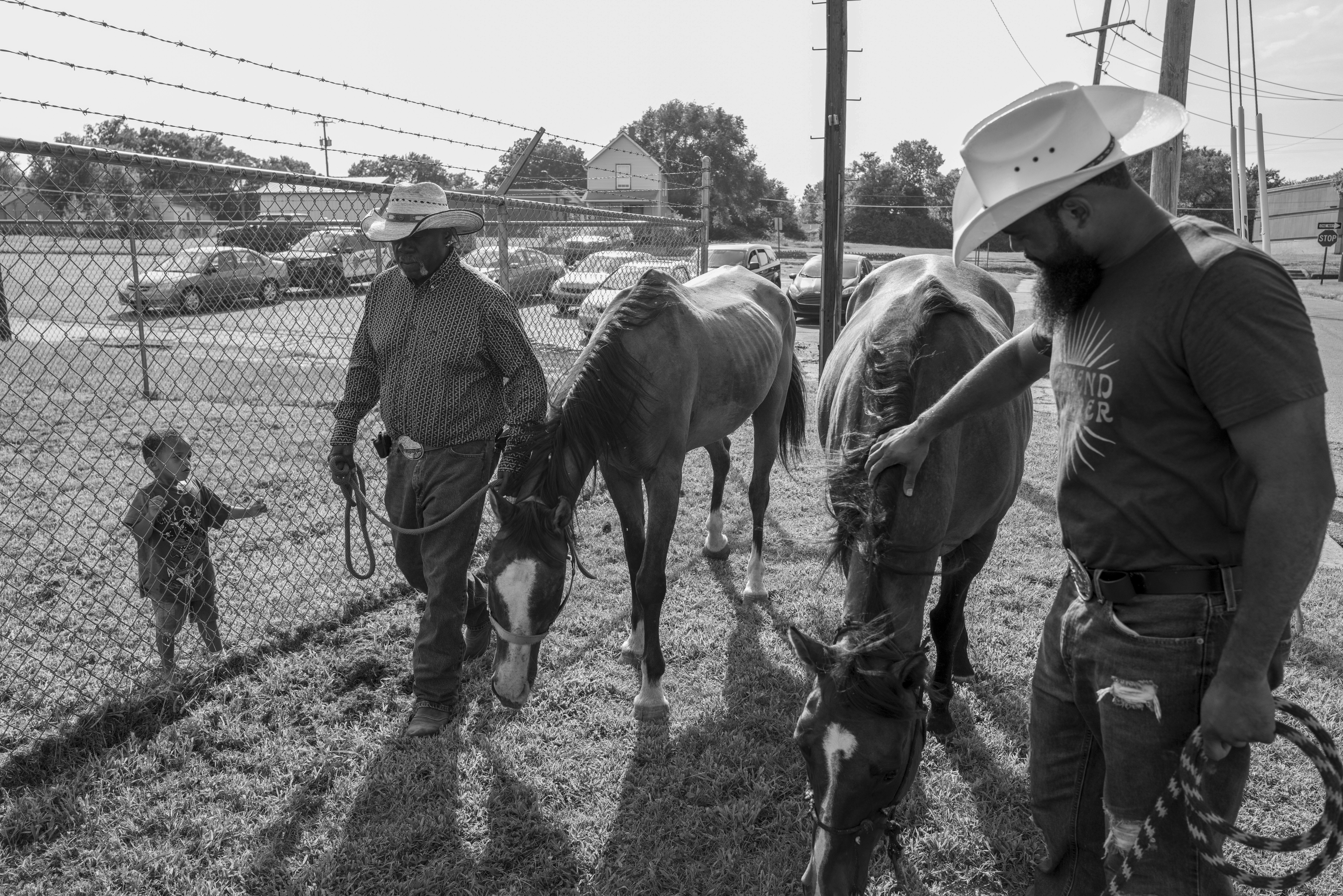 Black and white photograph showing two cowboys in cowboy hats leading horses through a ranch enclosure, with a small child reaching toward the fence in the foreground. Houses, utility poles, and chain-link fencing are visible in the background under an overcast sky.