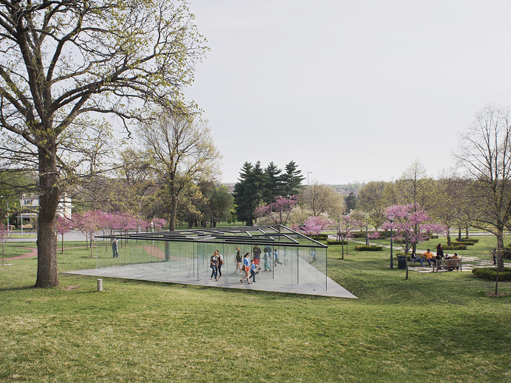 A contemporary pavilion with transparent glass walls and dark geometric roof panels sits in a manicured park setting. Several visitors walk through the structure while blooming magnolia trees in shades of pink dot the green landscape, with mature deciduous trees framing the composition.