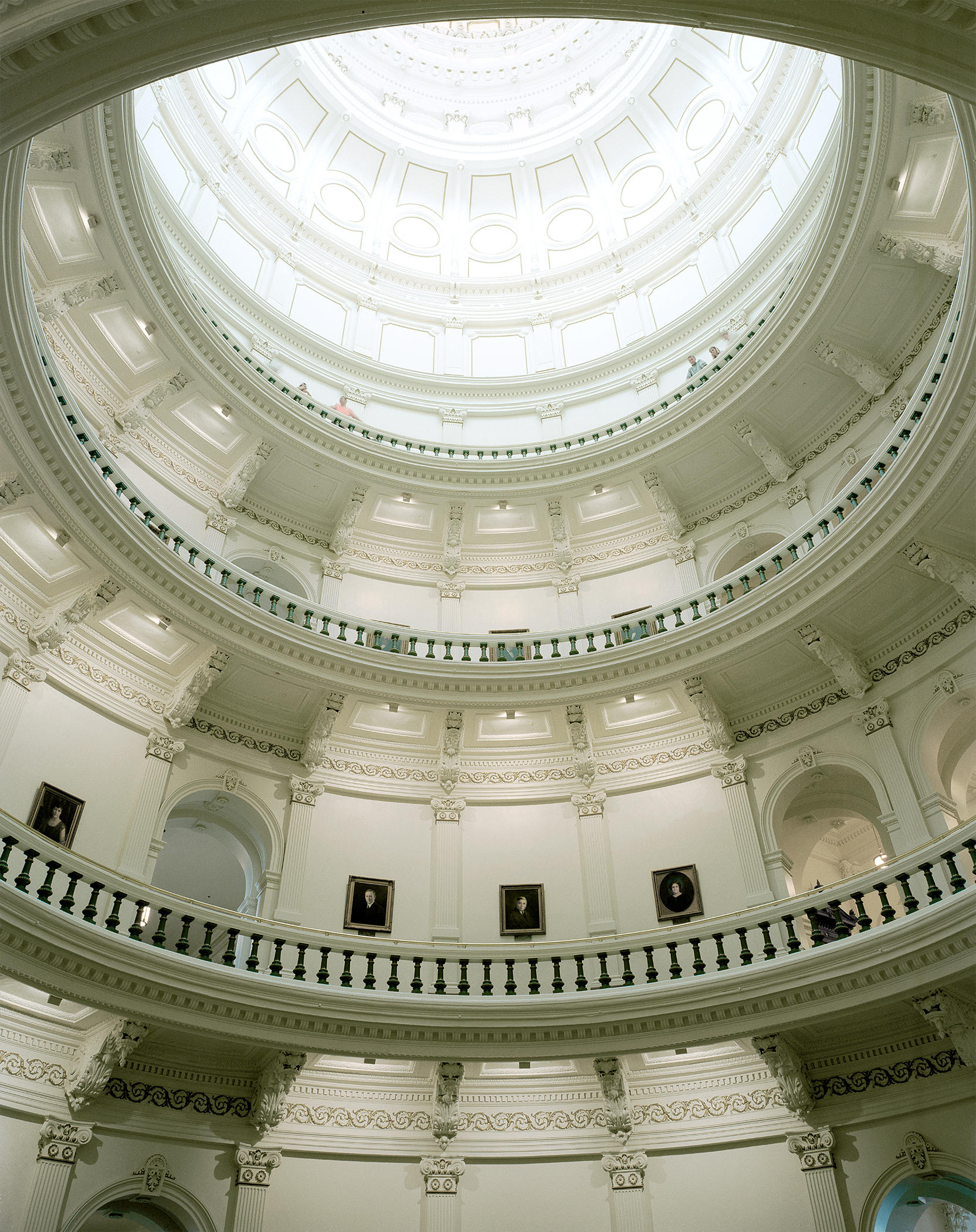 Symmetrical upward view of a grand neoclassical dome interior with concentric circular levels featuring ornate white plaster detailing, Corinthian capitals, and decorative moldings. Multiple tiers of arched openings and balustrades recede toward a central skylit oculus at the apex, with portrait paintings visible on the walls below.