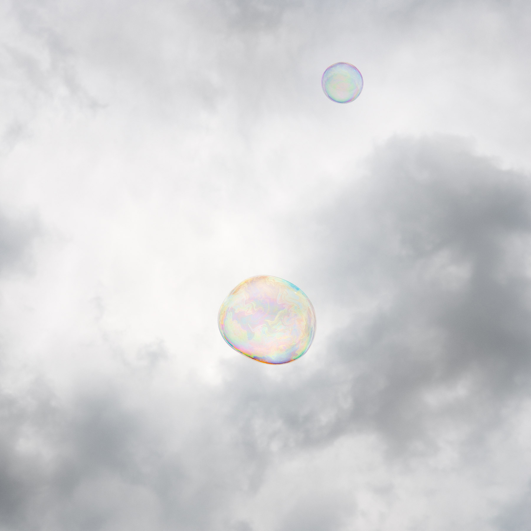 Two iridescent soap bubbles float against a soft, overcast sky with diffused clouds. The larger bubble in the lower center displays subtle rainbow hues across its translucent surface, while a smaller bubble hovers above it, creating a serene, minimalist composition.