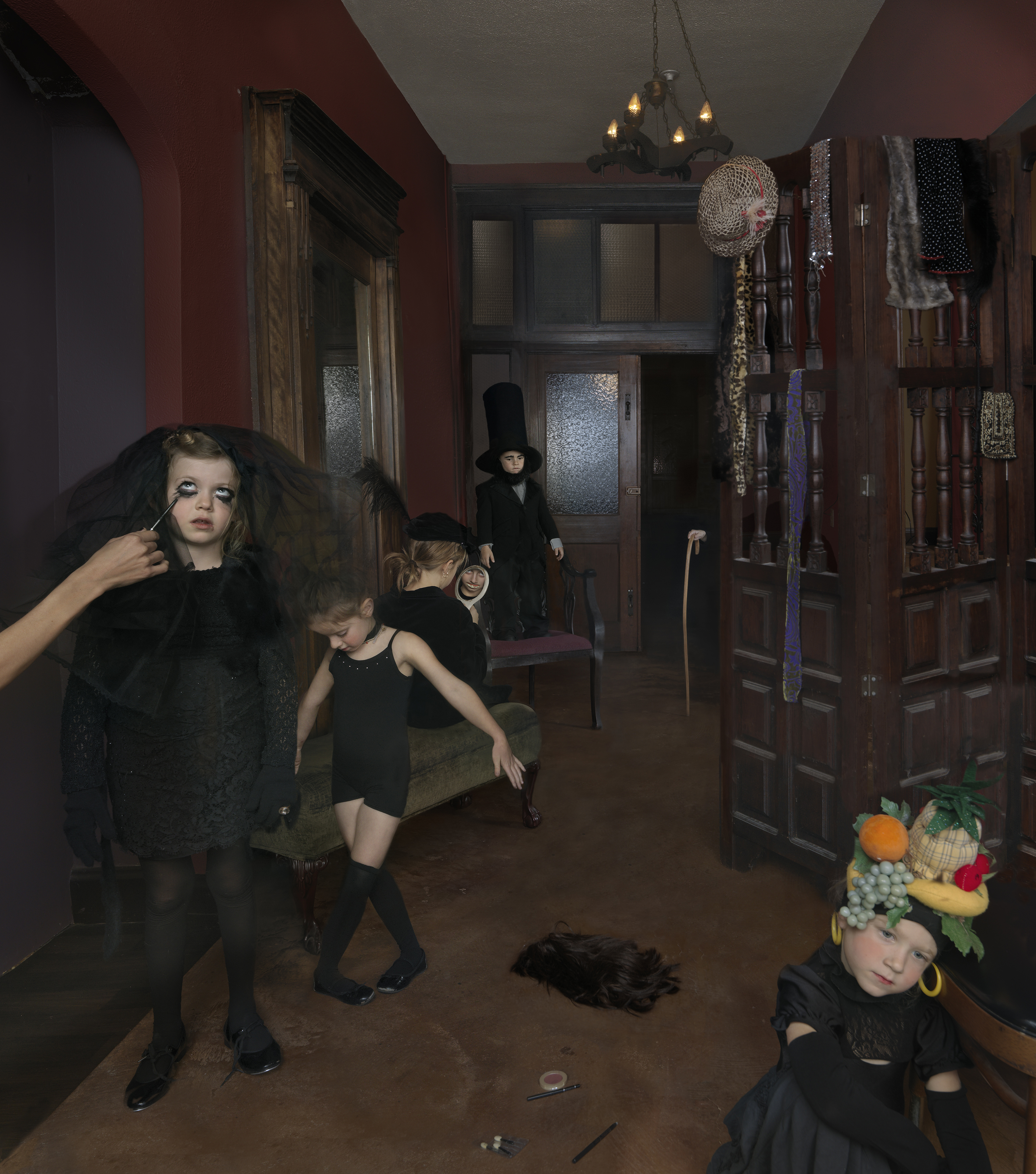 Children in dark clothing and theatrical makeup occupy a burgundy-walled room filled with wooden furniture and vintage accessories. A figure applies makeup to a child in the foreground while others pose throughout the space, one wearing an elaborate fruit and vegetable headdress, in a staged domestic scene with surreal, darkly whimsical undertones.
