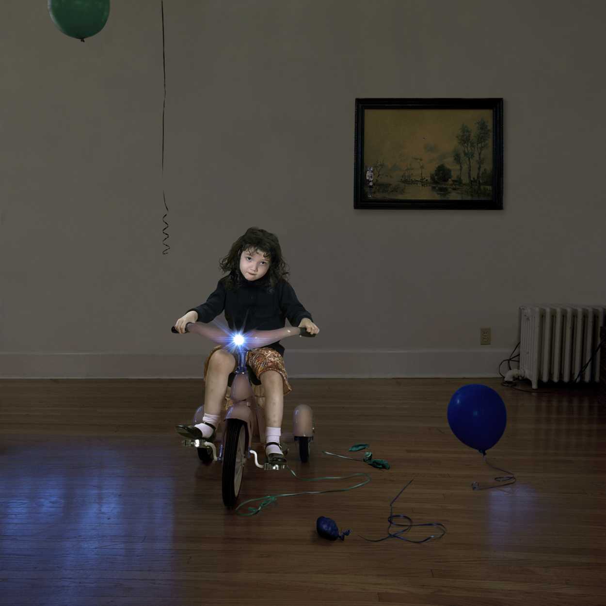 A child with curly dark hair rides a tricycle in a sparse interior space, holding illuminated blue light strands in their outstretched hands. A blue balloon tethered by a cord rests on the wooden floor nearby, while a framed landscape painting and radiator are visible on the walls behind.