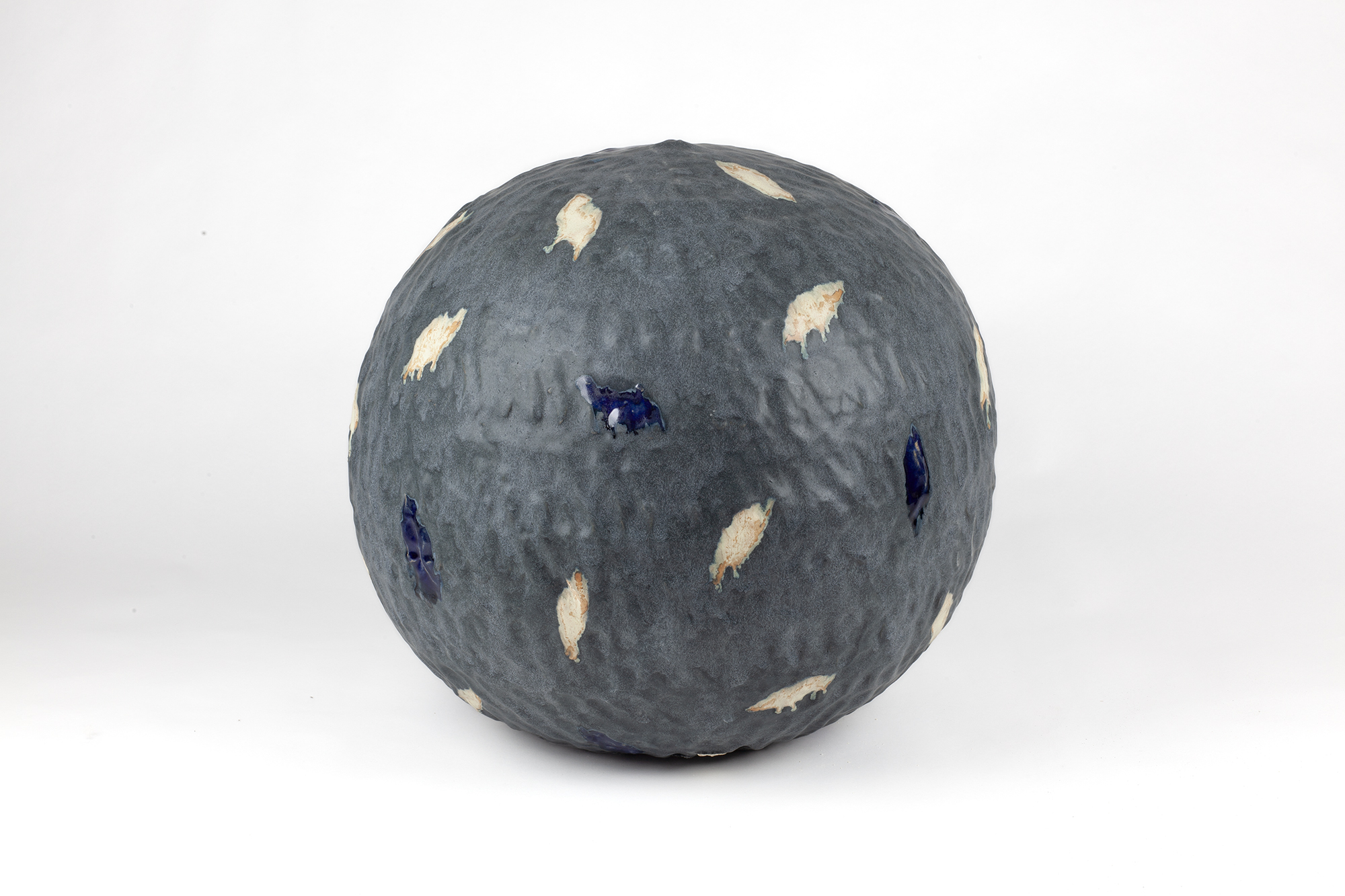 A spherical ceramic form with a textured grey surface scattered with cream-colored and deep blue leaf-shaped appliqués, displayed against a white background.