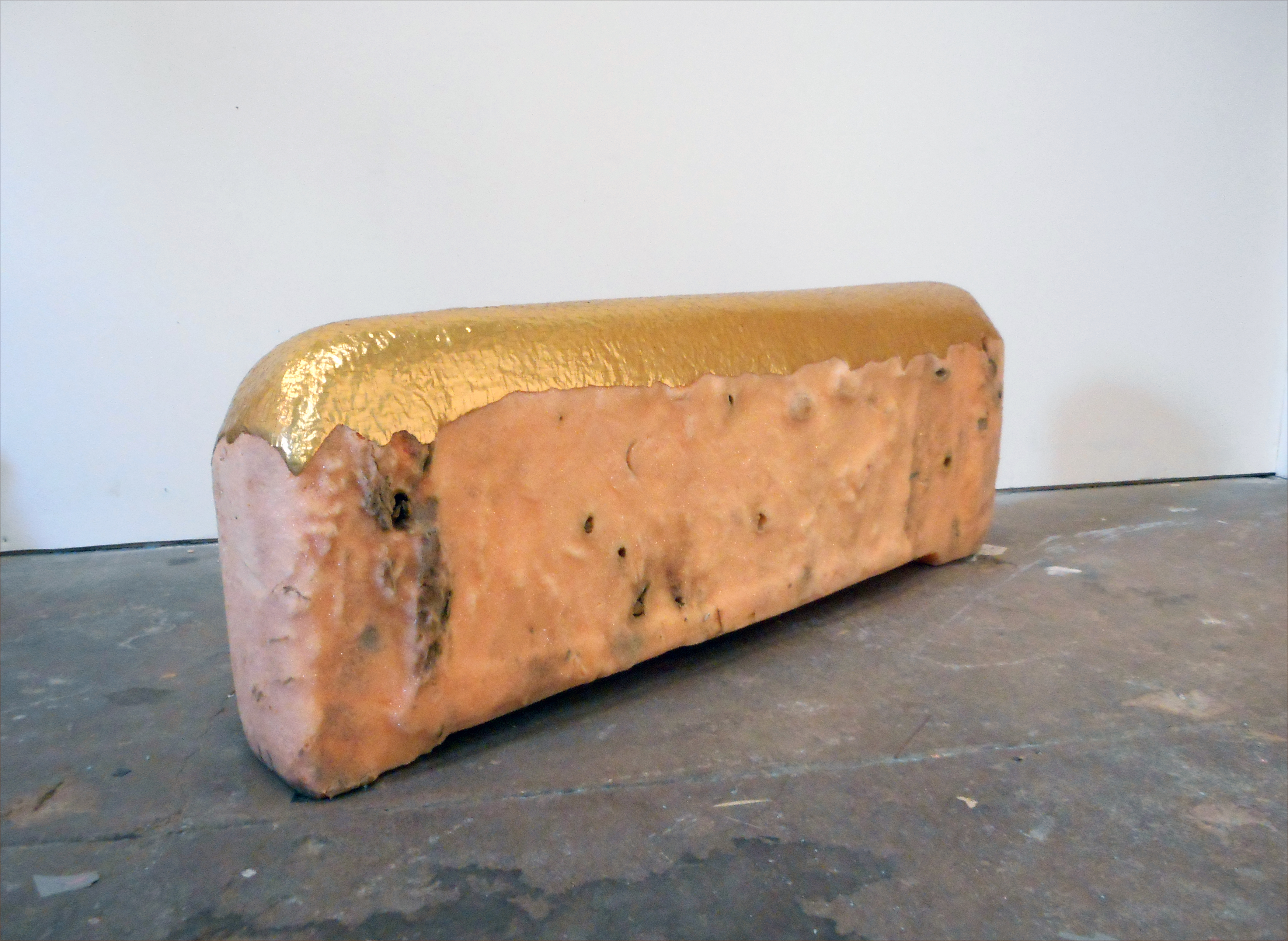 A cylindrical sculptural form with a salmon-pink body dotted with dark specks lies horizontally on a dark gallery floor. The top surface is wrapped in crinkled gold leaf, creating a stark contrast with the matte finish of the main body.