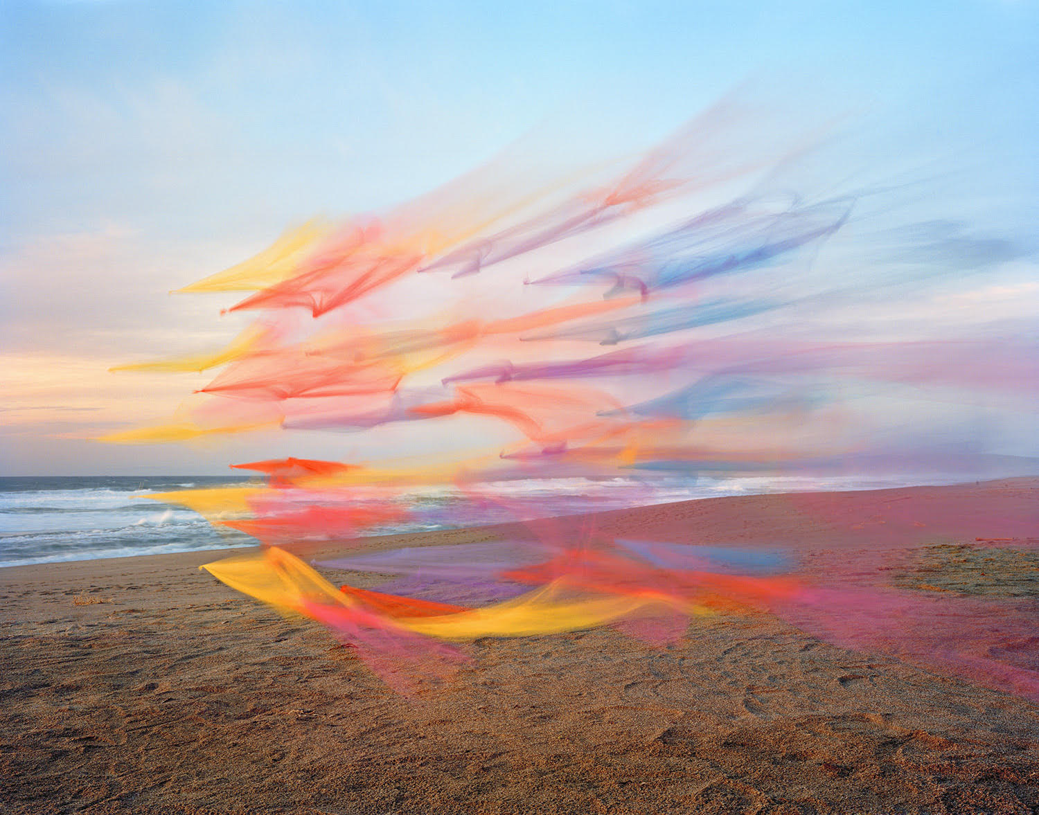 Colorful ribbons of yellow, orange, pink, and blue tulle fabric sweep dramatically across a sandy beach at sunset, creating flowing, gestural forms that contrast with the calm ocean and sky beyond.