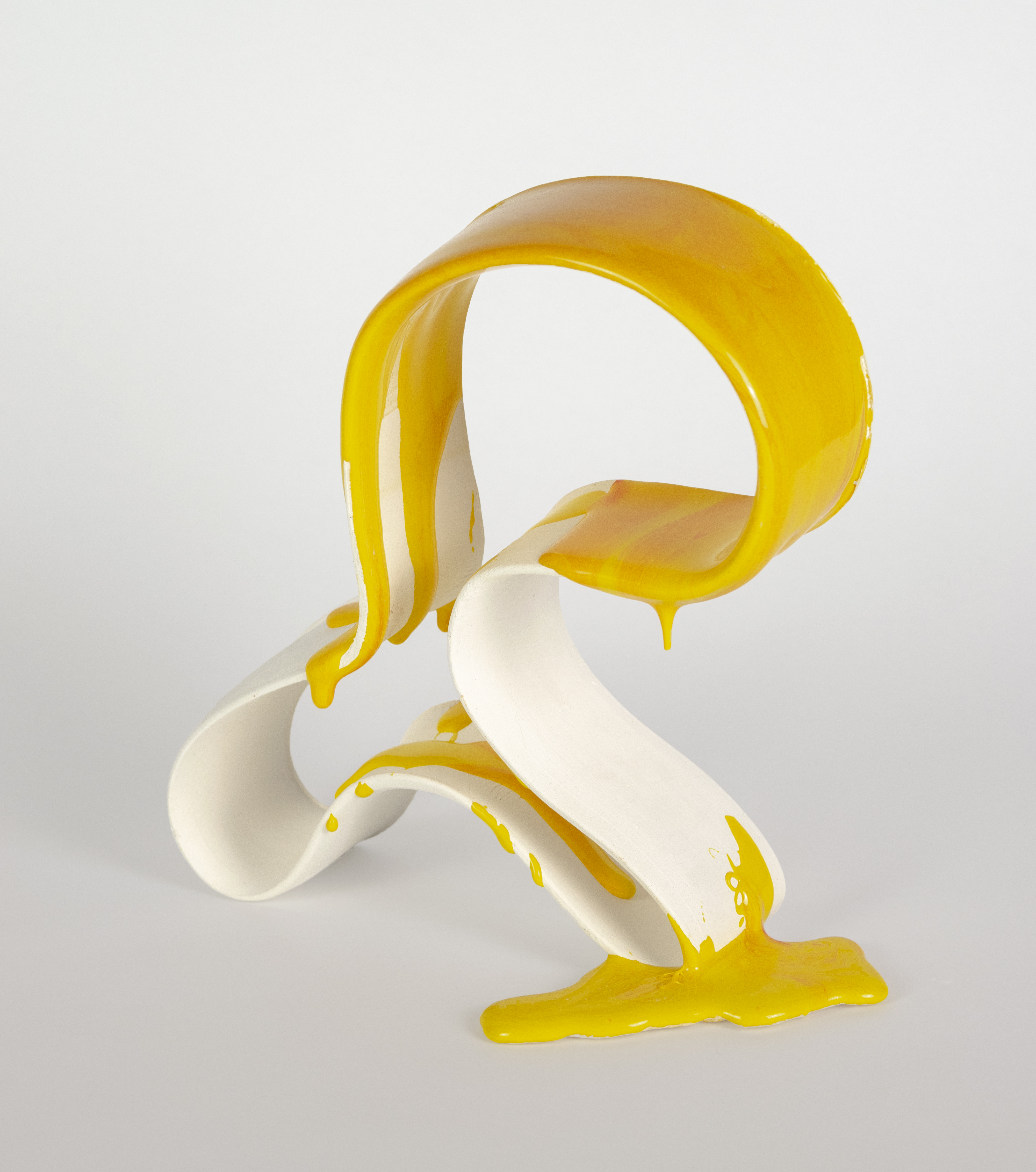 A sculptural arrangement of white and glossy yellow ceramic forms twists and curves organically, with curved bands and peeling shapes suggesting a stylized banana skin. Yellow liquid or glaze drips across the white surfaces, pooling at the base against a neutral gray background.