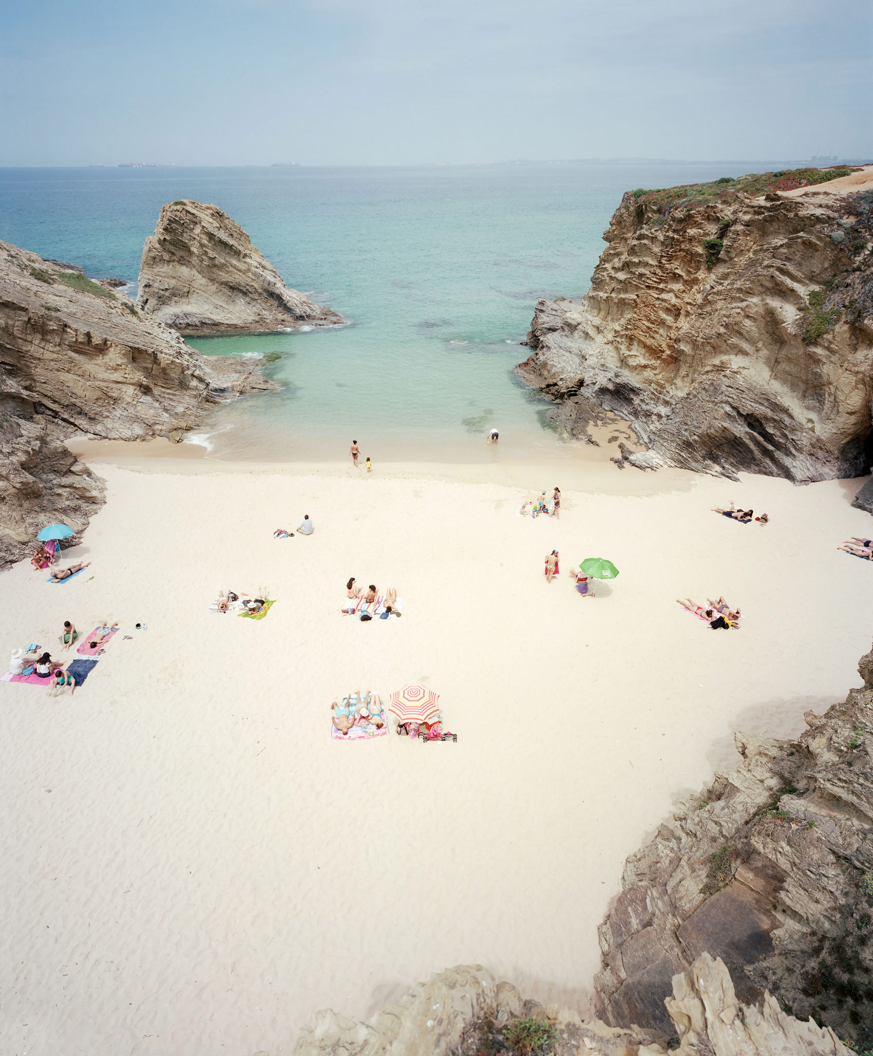 Aerial view of a secluded cove beach enclosed by dramatic white limestone cliffs, with turquoise and aquamarine waters. Scattered beachgoers in colorful swimwear and umbrellas dot the pale sand, creating vibrant pockets of color against the minimalist landscape.