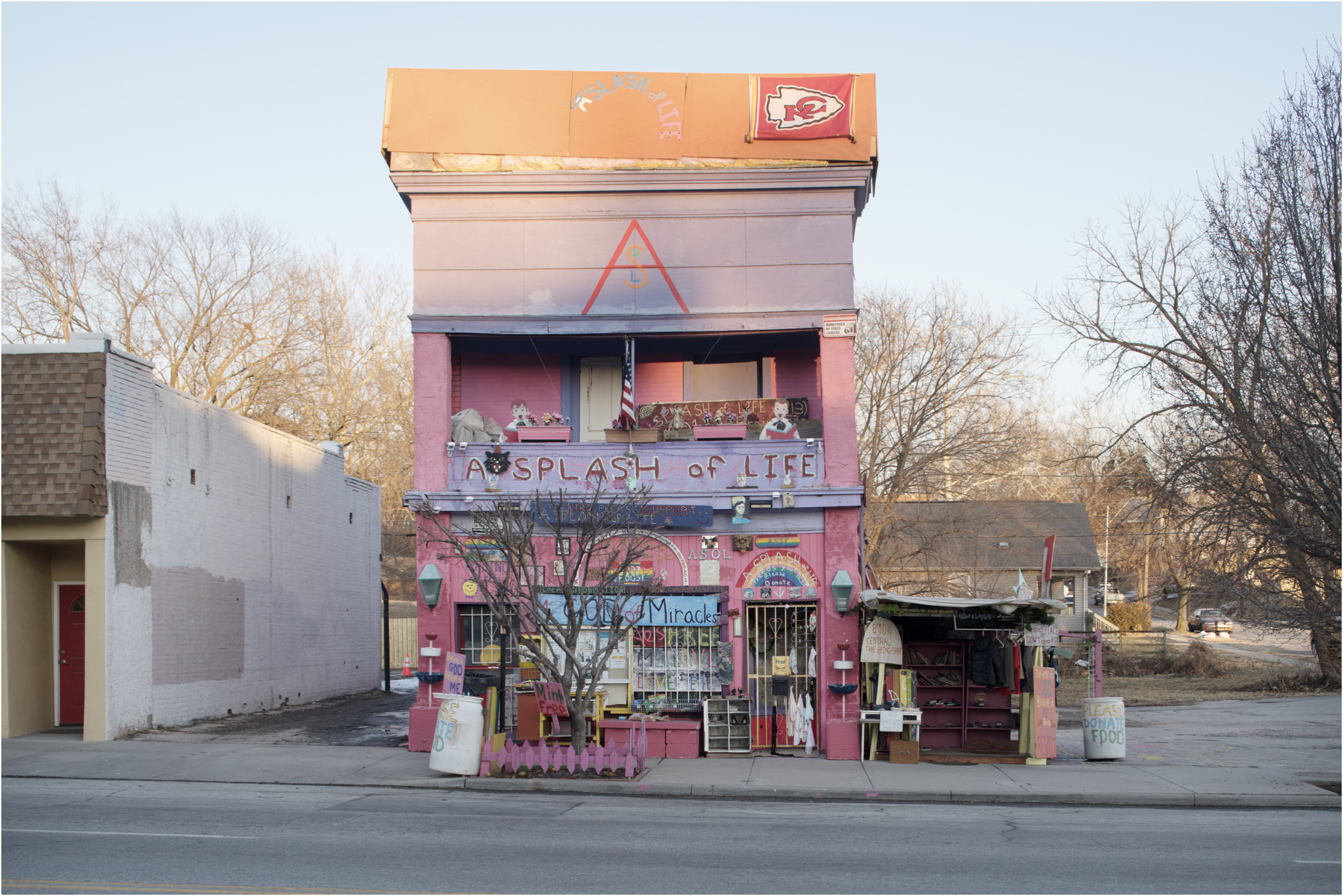 A bright pink, multi-story roadside structure with a Kansas City Chiefs logo on its roof sits abandoned along a flat, empty street. Bare trees and residential buildings flank the quirky edifice, which features weathered signage and scattered objects at its base, captured in cool daylight.