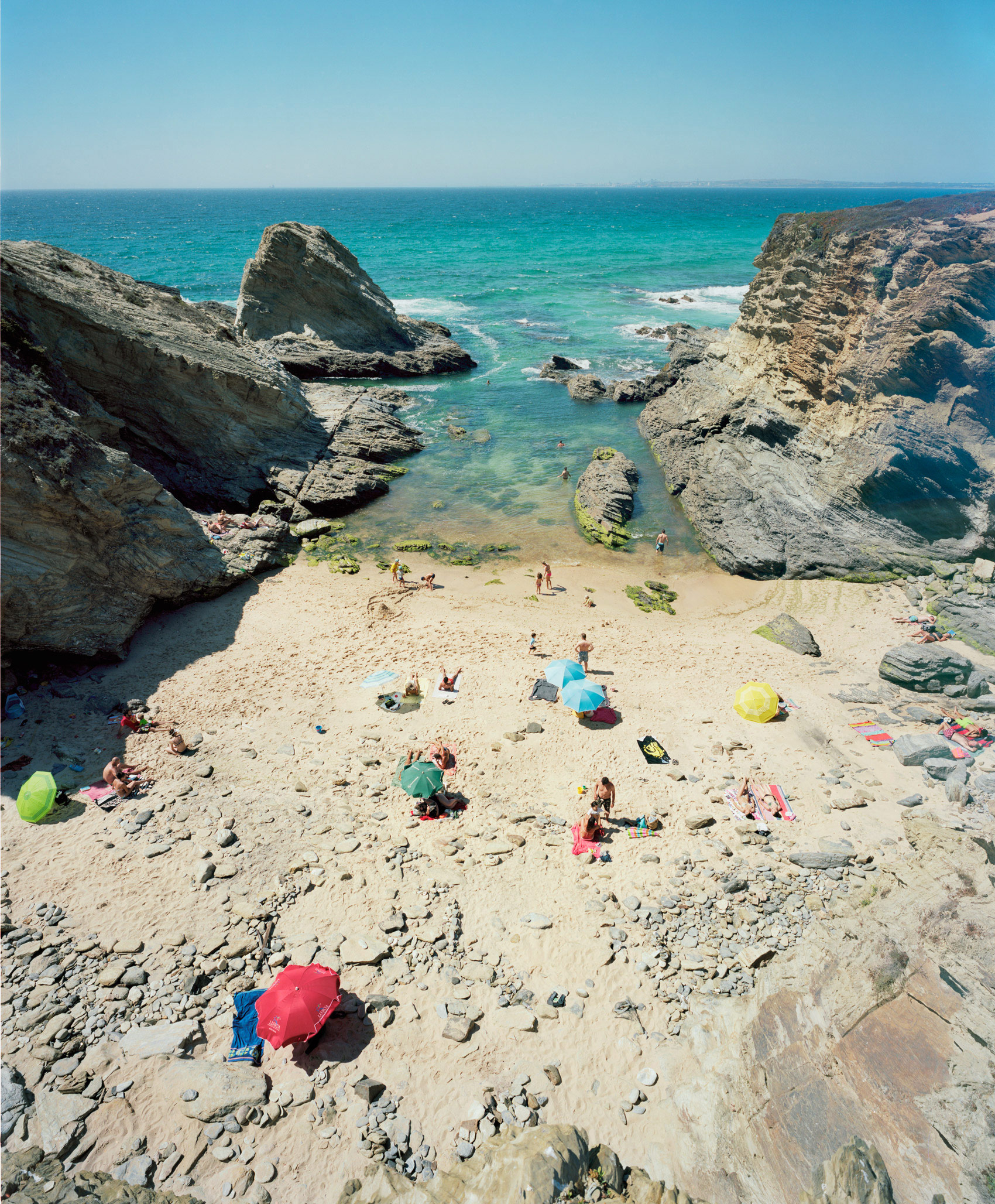 Aerial view of a secluded cove beach with turquoise water, dramatic rocky cliffs framing both sides, and scattered beachgoers with colorful umbrellas and towels dotting the pale sand. The composition captures the stark contrast between the vibrant blue sea, weathered gray stone formations, and the warm tones of the beach below.