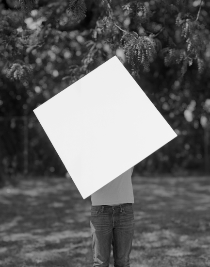 Black and white photograph of a figure standing in a natural landscape with overhanging foliage, their head replaced by a large white square that obscures their identity. The composition creates a stark geometric contrast between the blank rectangular form and the organic, textured surroundings.