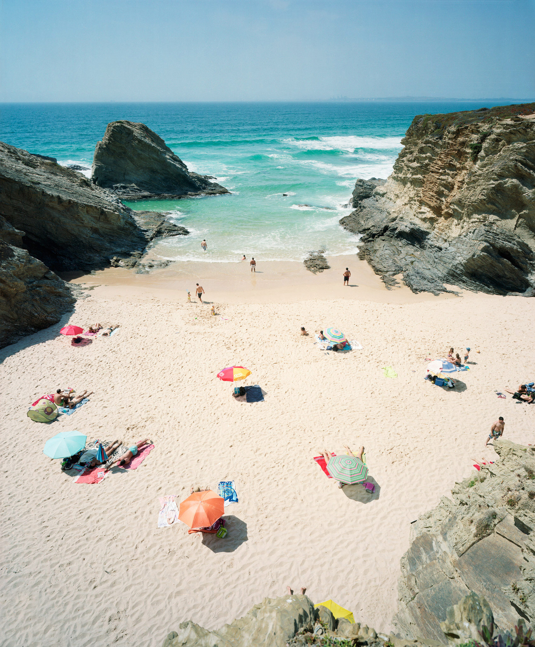 Aerial view of a secluded sandy beach nestled between dramatic rocky cliffs, with turquoise ocean waves and scattered beachgoers beneath colorful umbrellas and towels in shades of pink, orange, blue, and yellow.