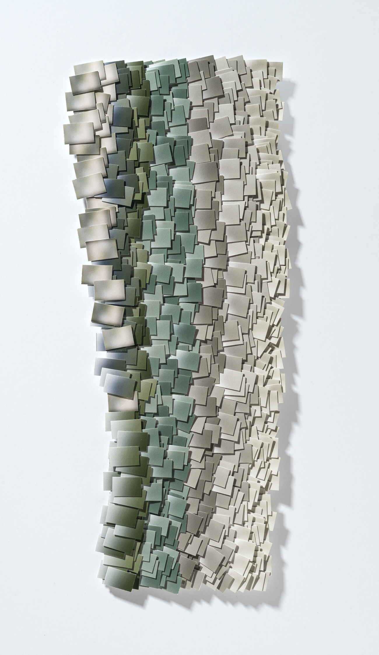 A vertical wall-mounted artwork composed of hundreds of hand-cut and layered rectangular paper fragments in graduated tones of sage green, taupe, and cream, creating a three-dimensional textured surface with pronounced shadows that suggest a pixelated or topographic landscape.
