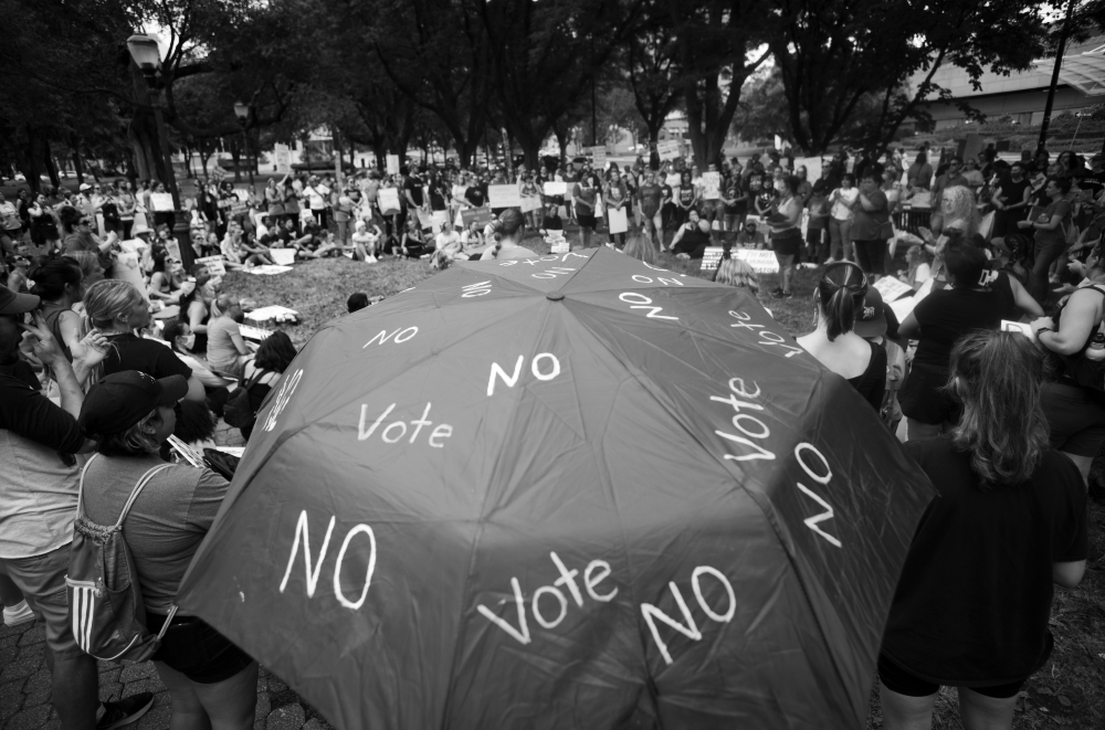 Black and white photograph of a large outdoor gathering in a park, with hundreds of people surrounding a prominent dark umbrella inscribed with repeated text reading "NO" and "vote." Tall trees frame the scene, and participants hold signs while seated and standing around the central umbrella, documenting a political protest or demonstration.
