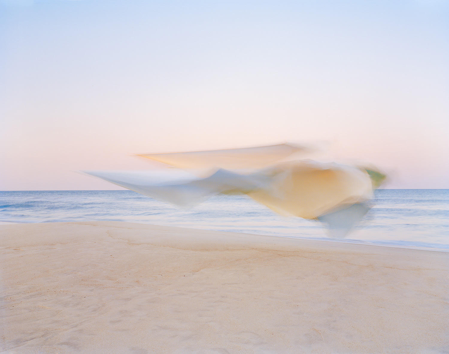 Soft-focus photograph of cream and pale blue fabric billowing in motion above a beach shoreline, with calm ocean and clear sky creating a serene, dreamlike composition.