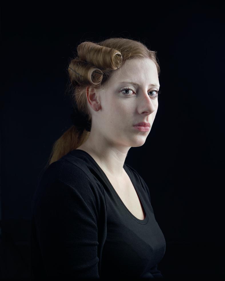 Portrait of a woman in profile against a black background, wearing a black long-sleeved top. Her hair is styled in vintage waves and pinned up on one side, with a ponytail visible. Her gaze is directed downward and to the side, creating a contemplative mood. The lighting is soft and controlled, emphasizing the delicate features and pale complexion of her face.