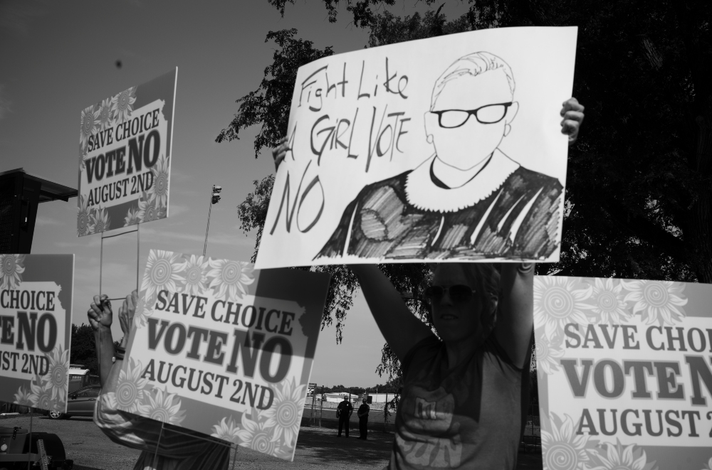 Black and white photograph of a protest scene with multiple handmade signs held aloft. Prominent signs read "SAVE CHOICE VOTE NO AUGUST 2ND" with decorative floral patterns, while another large sign features a portrait sketch of a person wearing glasses alongside the text "FIGHT LIKE GIRL VOTE NO." Trees and sky are visible in the background, with several protesters partially visible behind the signs.