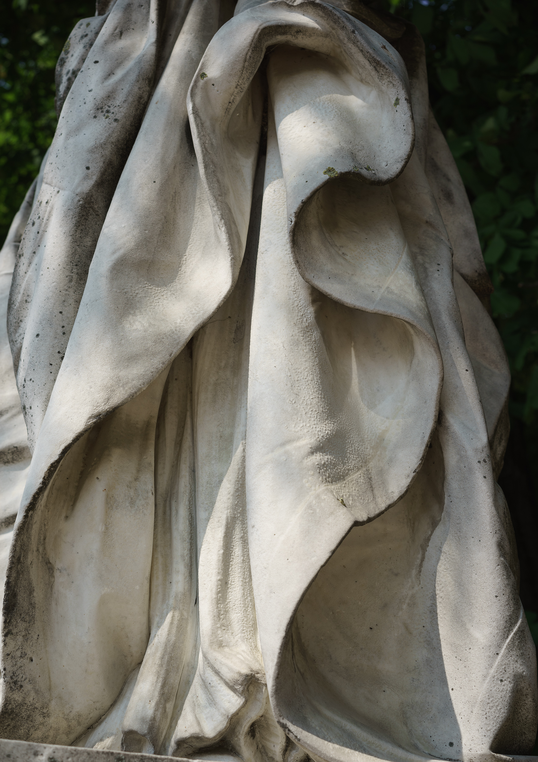 Close-up detail of a stone sculptural form with flowing, draped fabric carved in relief. The pale limestone or marble surface catches strong directional light, creating deep shadows within the undulating folds and hollow cavities, emphasizing the texture and dimensionality of the carved drapery against a softly blurred green background.
