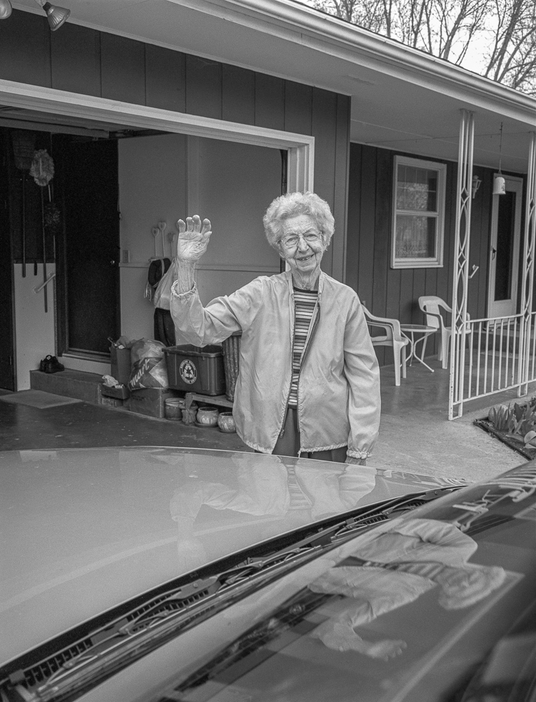 Black and white photograph of an elderly woman with white hair and glasses waving from the hood of a car parked in front of a modest single-story house with a carport. She wears a light-colored jacket over a striped shirt and smiles at the camera while leaning against the vehicle's windshield.