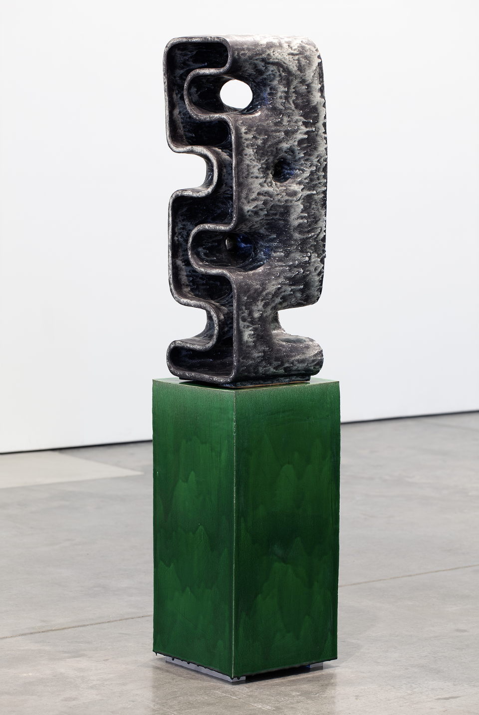 Dark stoneware sculpture with undulating wavy edges and a circular opening, mounted on a bright green cubic pedestal. The textured black ceramic form stands vertically with an organic, gate-like silhouette against a white gallery wall.