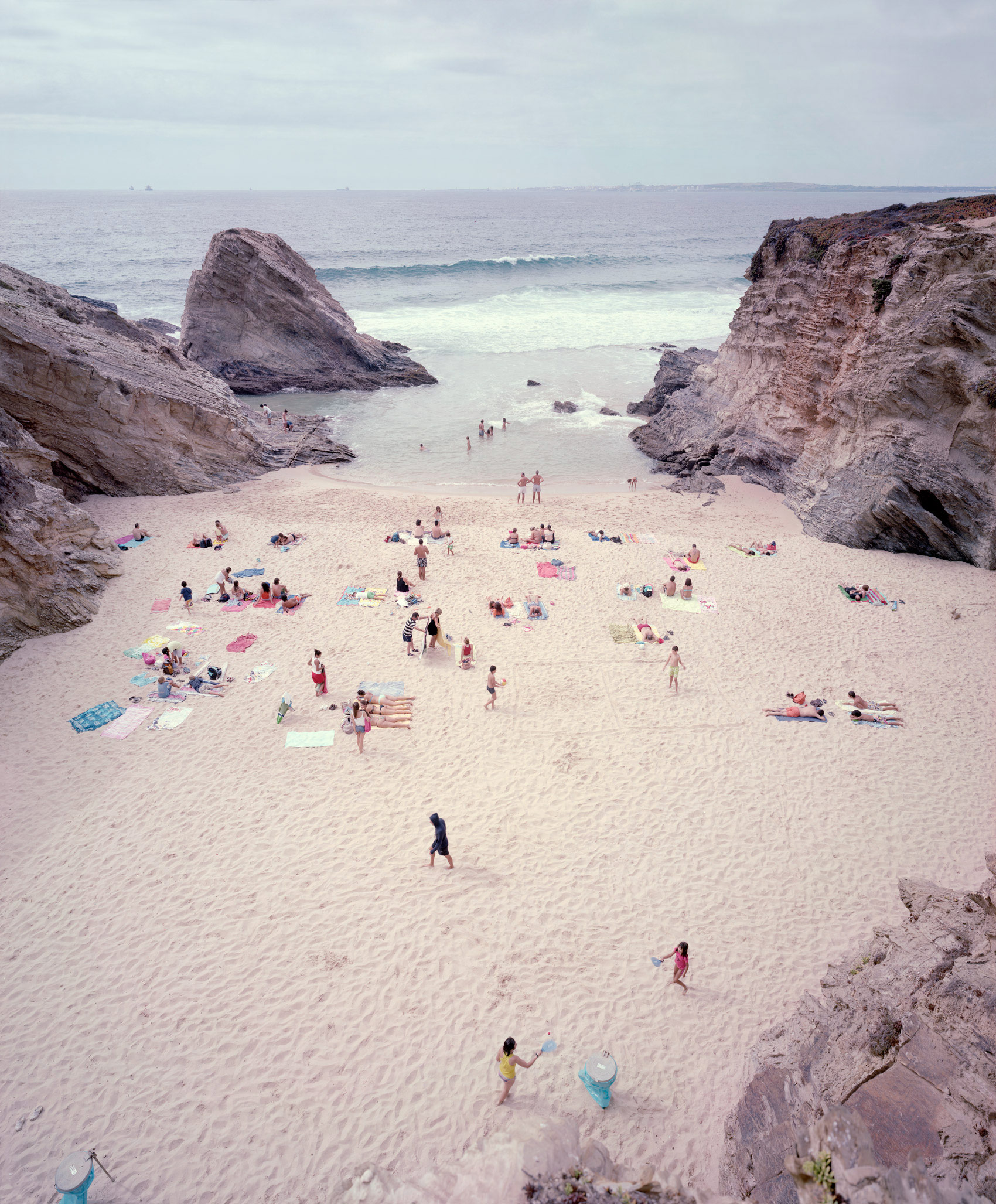 Aerial view of a crowded beach nestled between two large rocky outcroppings, with beachgoers scattered across pale sand in colorful swimwear and towels. Calm turquoise waters and distant horizon visible beyond the beach, with a few swimmers wading in the shallow sea.