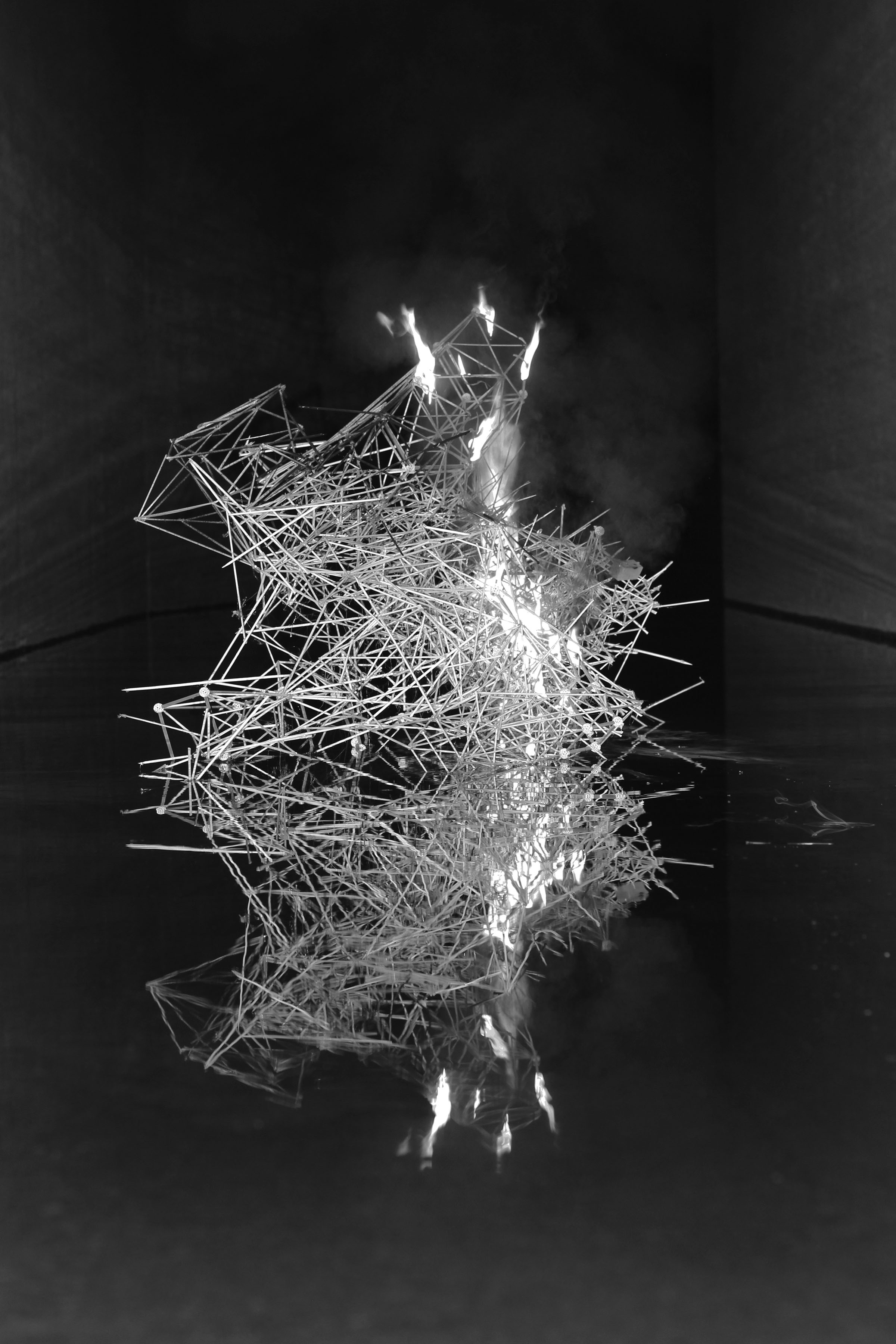A luminous white geometric wireframe structure composed of intersecting lines and triangular forms rises against a black background, creating a three-dimensional architectural abstraction with glowing highlights that suggest internal light or reflective surfaces.