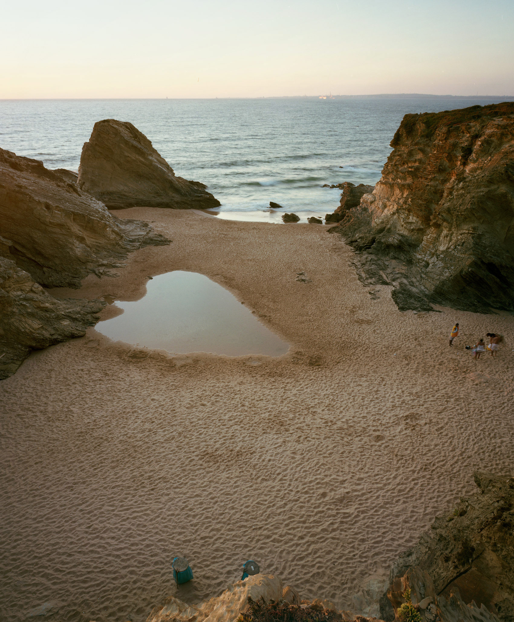 A secluded sandy beach nestled between dramatic rocky cliffs, with a shallow tidal pool reflecting the pale sky in the foreground. A few figures stand in the shallow waters and on the beach, dwarfed by the towering geological formations that frame the composition, under a soft, muted twilight sky.