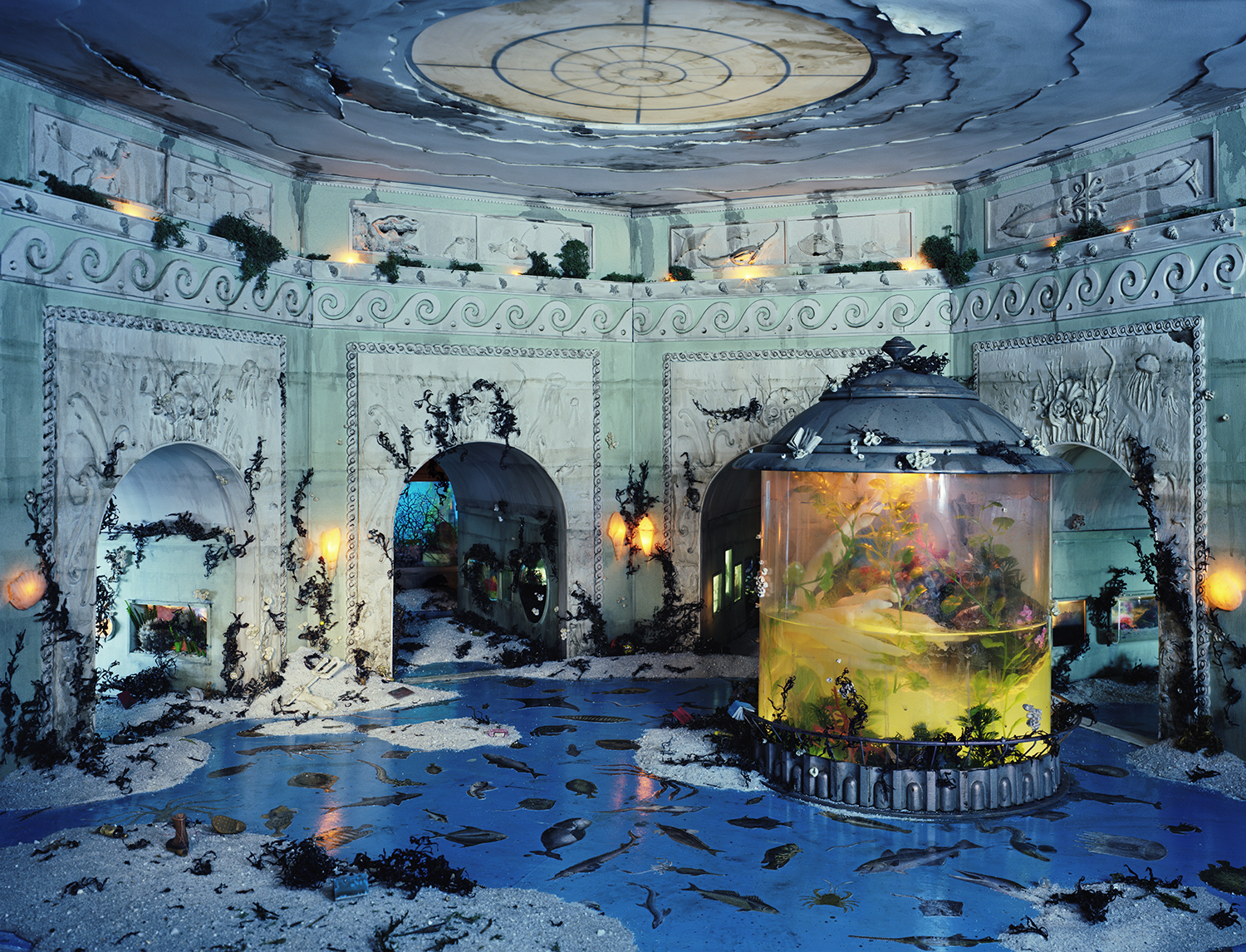 A grand circular room with turquoise walls and ornate neoclassical detailing features large arched doorways adorned with climbing vines. The floor is flooded with deep blue water scattered with fish and dark debris, while a central cylindrical aquarium tank glows with yellow-green liquid and aquatic life. Warm sconce lighting and an elaborate ceiling with a geometric skylight complete the deteriorated, fantastical interior.
