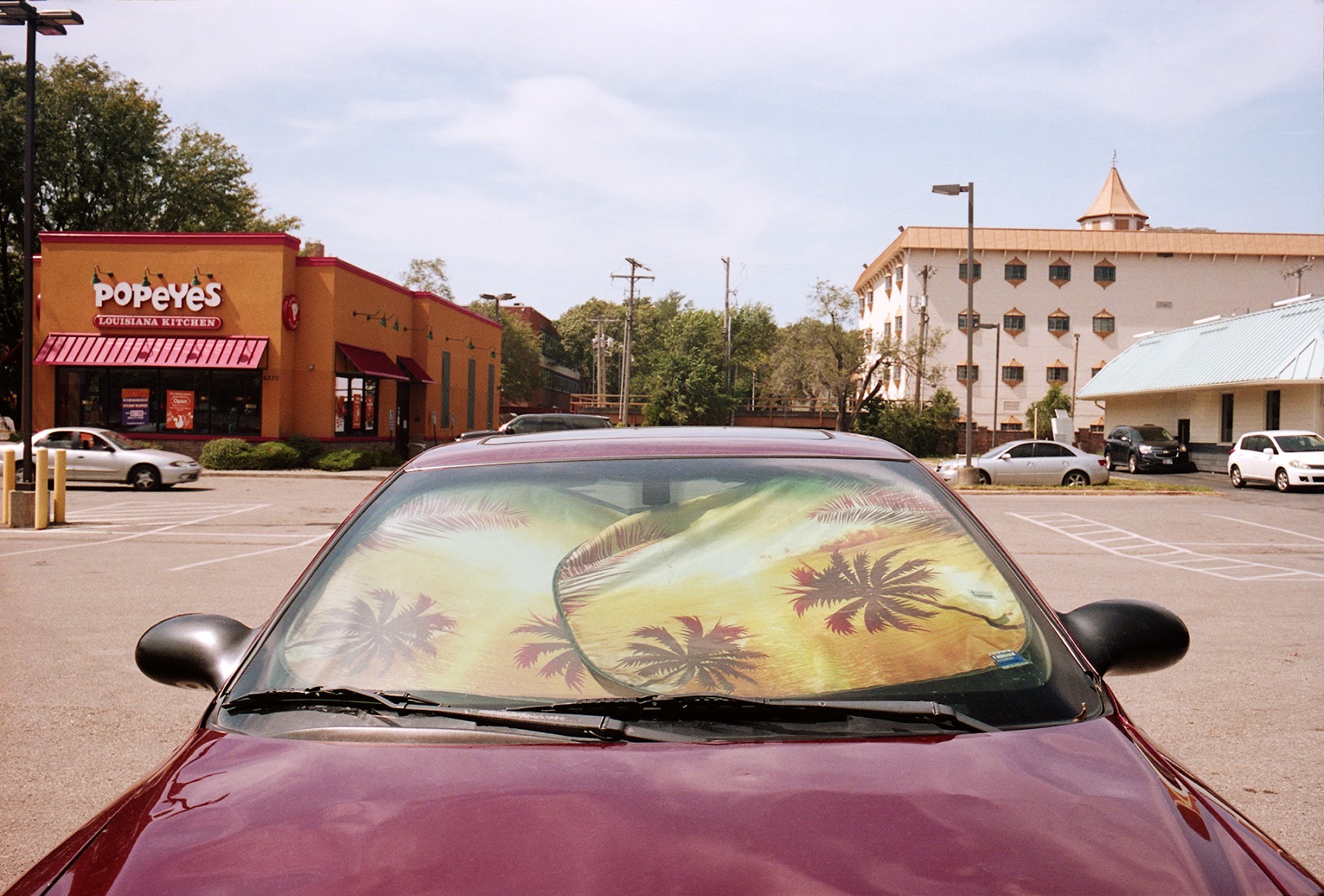 A maroon car parked in a commercial plaza displays a sunshade across its windshield featuring a tropical landscape with palm trees rendered in warm yellows and greens. Behind the vehicle, a Popeyes Louisiana Kitchen restaurant and multi-story building are visible under a partly cloudy sky.