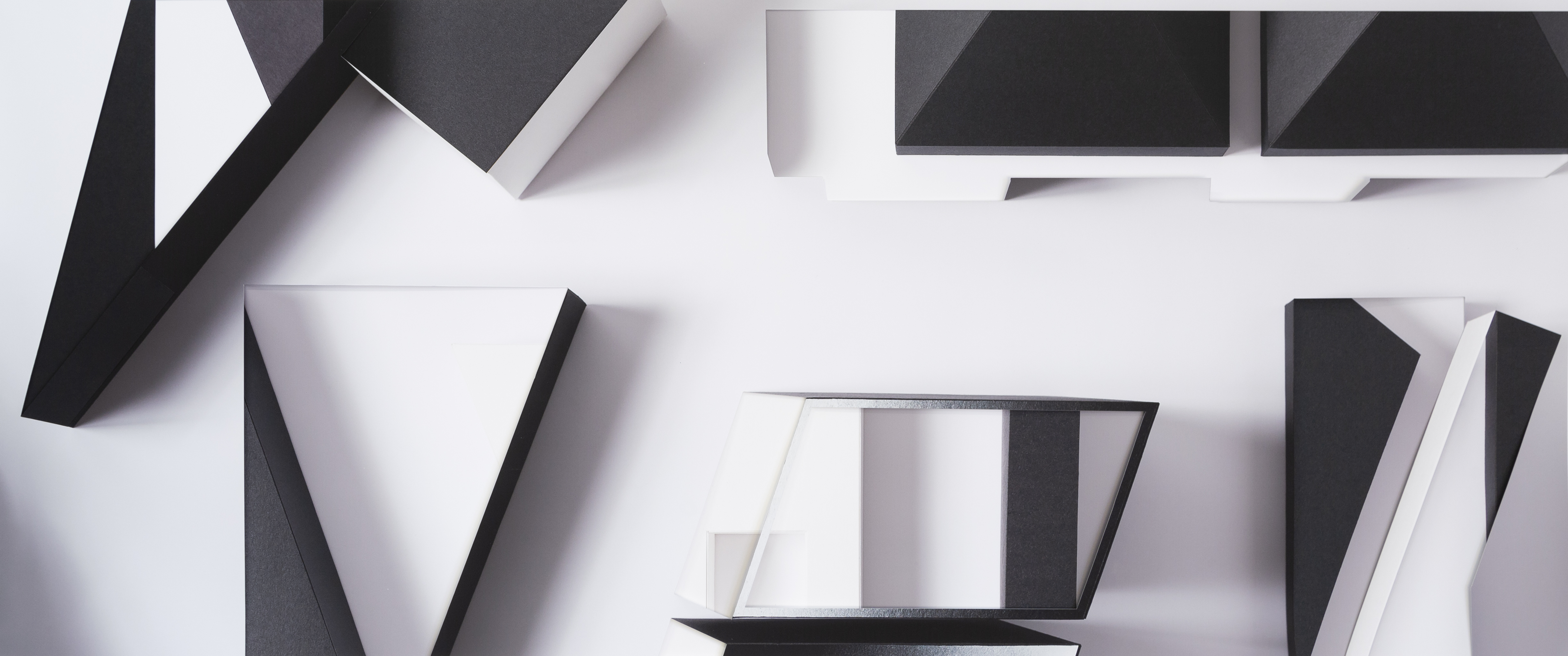 Geometric abstract composition of layered, fragmented forms in black, white, and gray tones arranged across a light background. Sharp angular shapes and rectangular planes overlap and cast shadows, creating depth and visual tension through a deconstructed, collage-like arrangement.