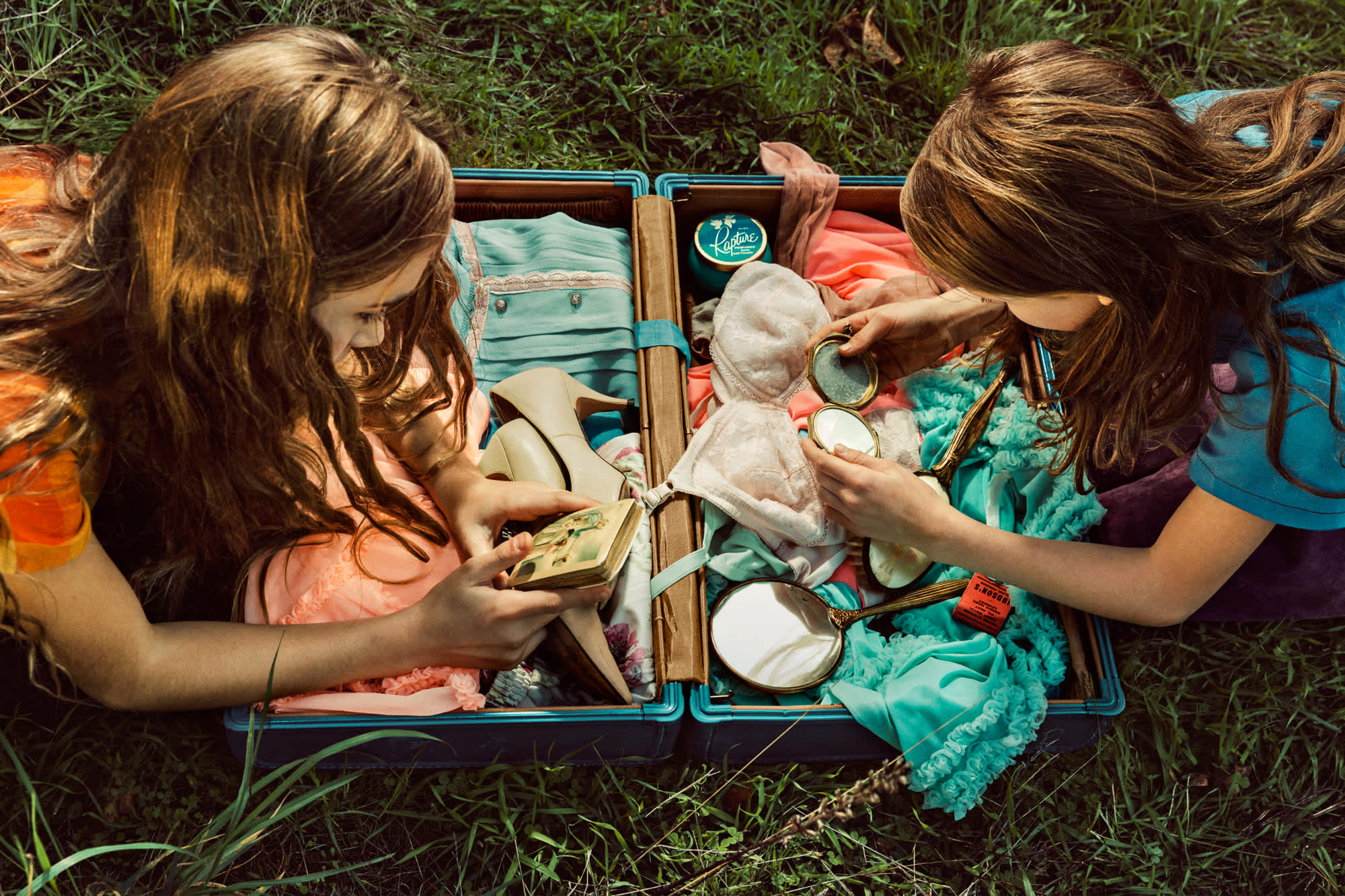 Two girls sit on grass beside an open turquoise and coral-colored suitcase, examining its contents of clothing and mirror accessories. Shot from above, the photograph captures warm sunlight casting shadows across their brown hair and the vibrant textiles arranged within the vintage luggage.