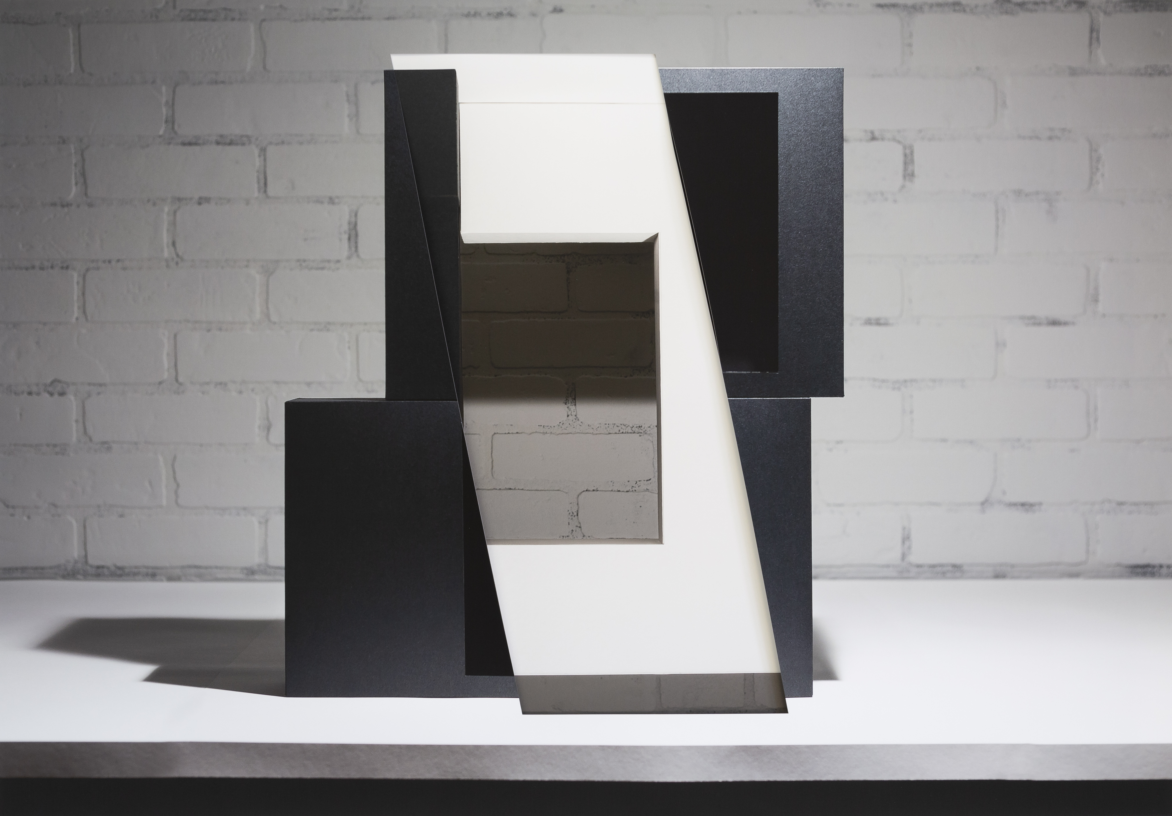 Abstract geometric composition featuring layered rectangular forms in black, white, and gray tones arranged to create depth and dimensional space. The collaged and layered construction reveals cut and overlapping planes that cast shadows against a white brick wall backdrop.