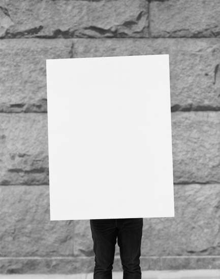 A figure in dark clothing holds a large blank white rectangle in front of their body, obscuring their head and torso against a gray brick wall. Only their lower legs and feet are visible below the white sheet.