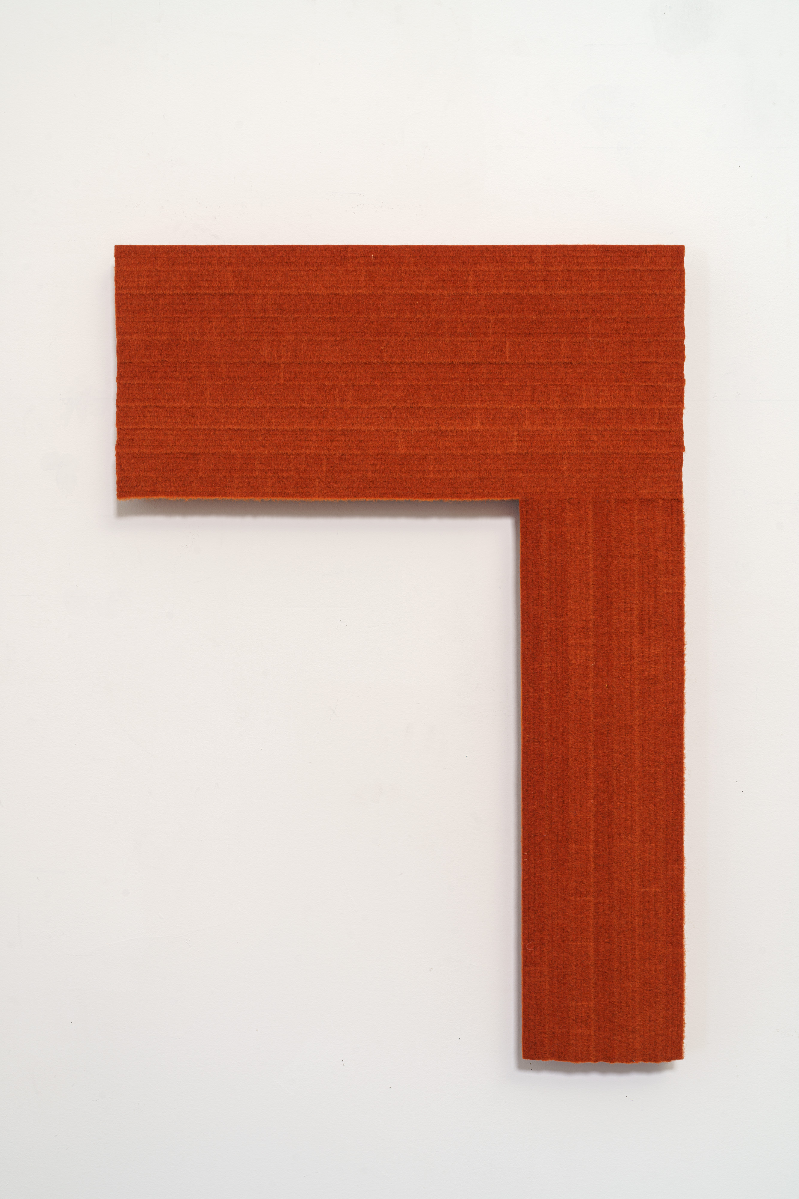 A bold rust-orange geometric form in the shape of a right angle or inverted "L" mounted on a white wall. The piece has a textured, matte fabric surface with subtle color variation.