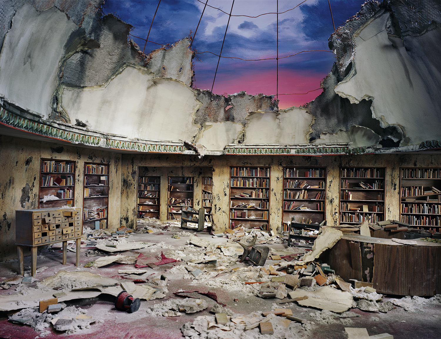 A devastated library interior with rows of bookshelves lining crumbling walls, their spines still visible among scattered debris and rubble covering the floor. The ceiling is partially destroyed, framing a dramatic twilight sky in shades of blue and pink above the scene of decay.