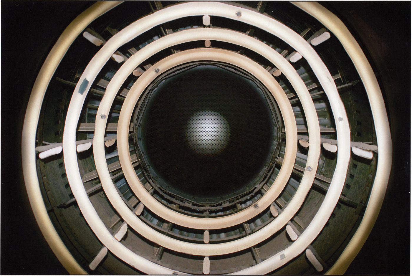 Concentric circles of cream and white rings with metal fixtures frame a dark central aperture, creating a geometric spiral effect. The composition suggests looking down into a cylindrical industrial or architectural structure, with alternating bands of light and shadow receding toward a barely visible illuminated center.