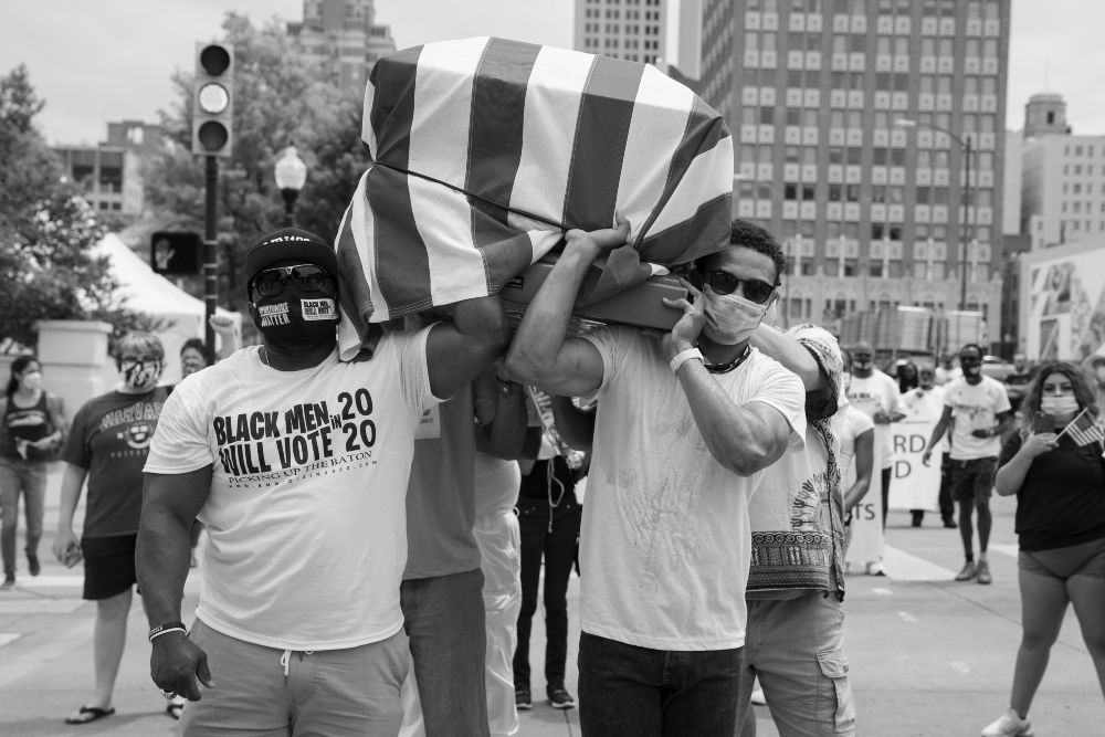 Black and white photograph of protesters in an urban plaza holding a large striped flag aloft. Two men in the foreground wear white t-shirts, one reading "Black Men Vote 20," while surrounding figures hold signs and gather in demonstration. Tall buildings frame the scene against a cloudy sky.