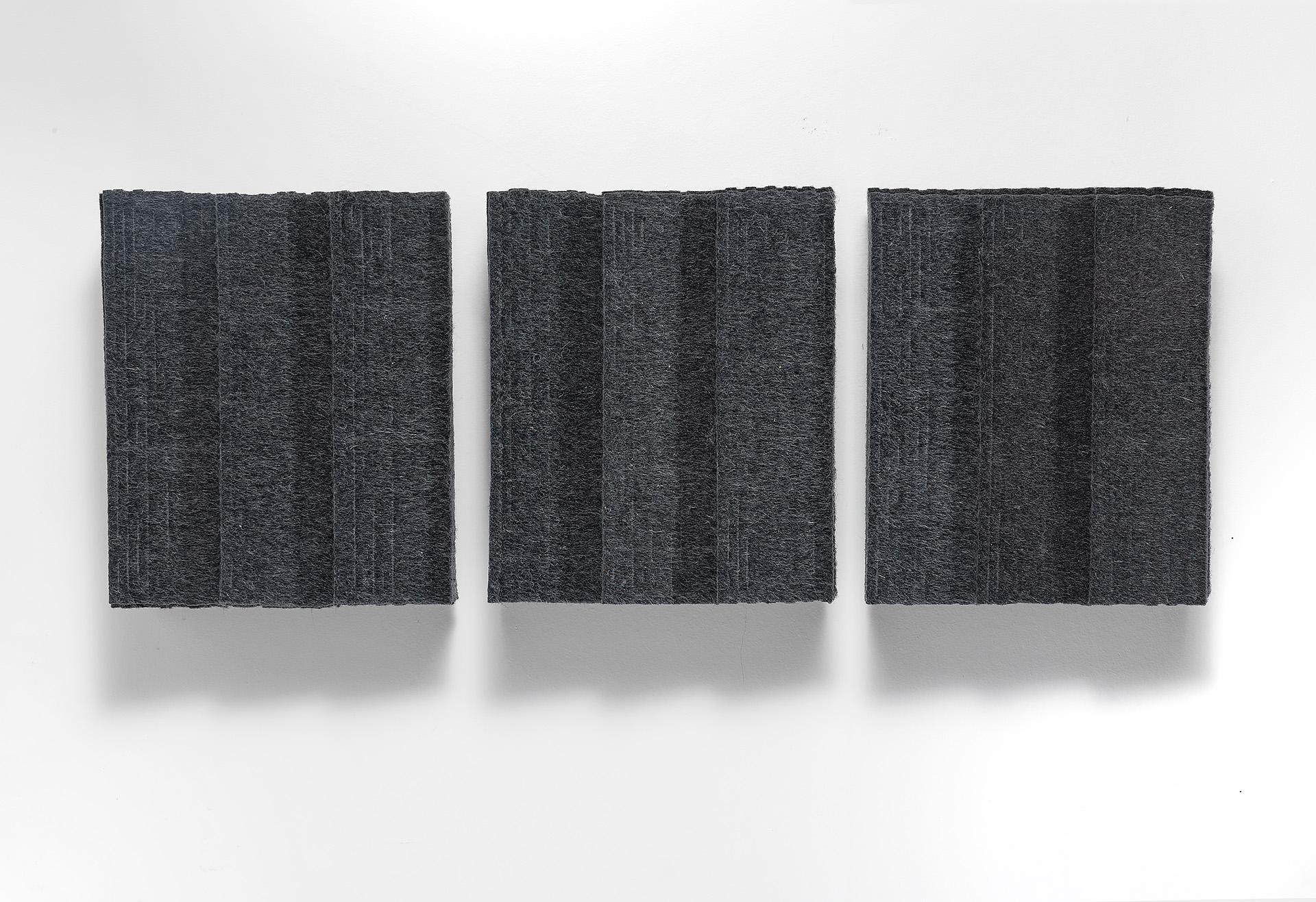 Three dark gray textile panels arranged horizontally against a white wall, each featuring vertical pleated or folded sections that create subtle linear patterns across their surfaces. The panels cast soft shadows, emphasizing their three-dimensional, sculptural quality.
