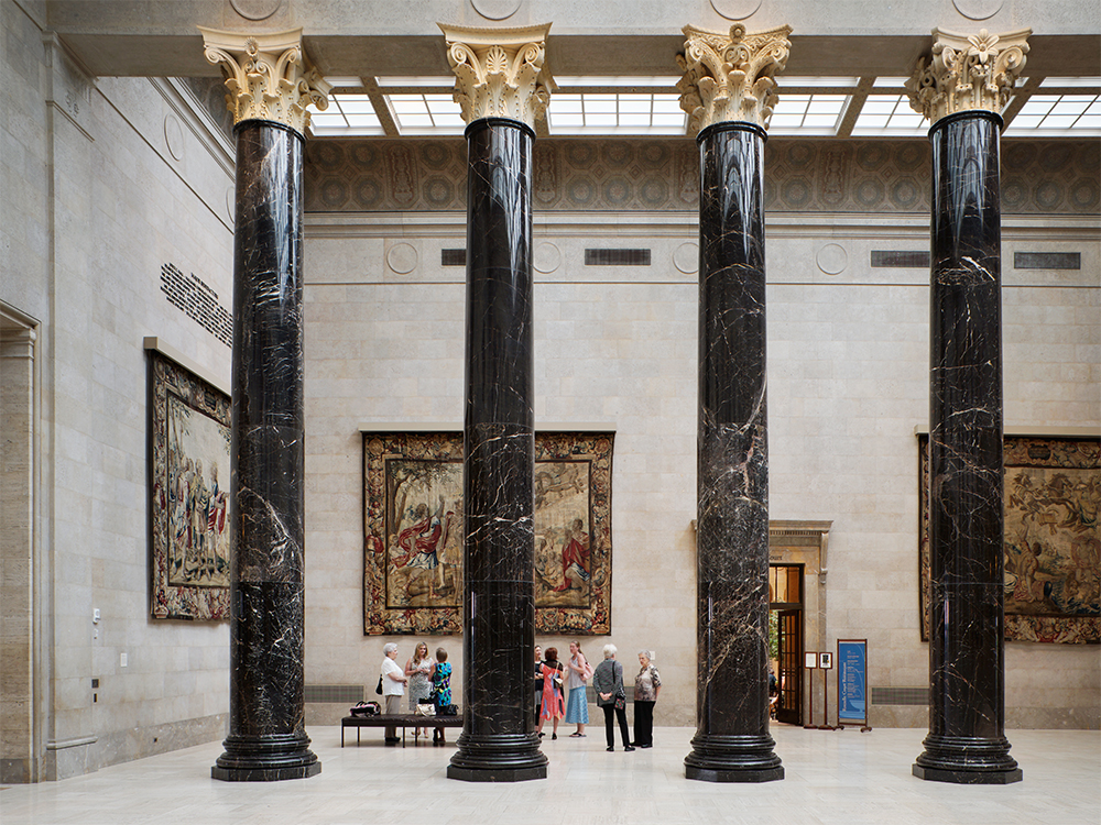A grand neoclassical interior featuring four towering black marble columns with ornate gilded Corinthian capitals supporting a coffered ceiling with skylights. Visitors stand on a polished stone floor beneath the columns, while tapestries depicting classical scenes hang on cream-colored walls between the columns. The composition emphasizes symmetry, scale, and the interplay of dark materials against light architectural surfaces.