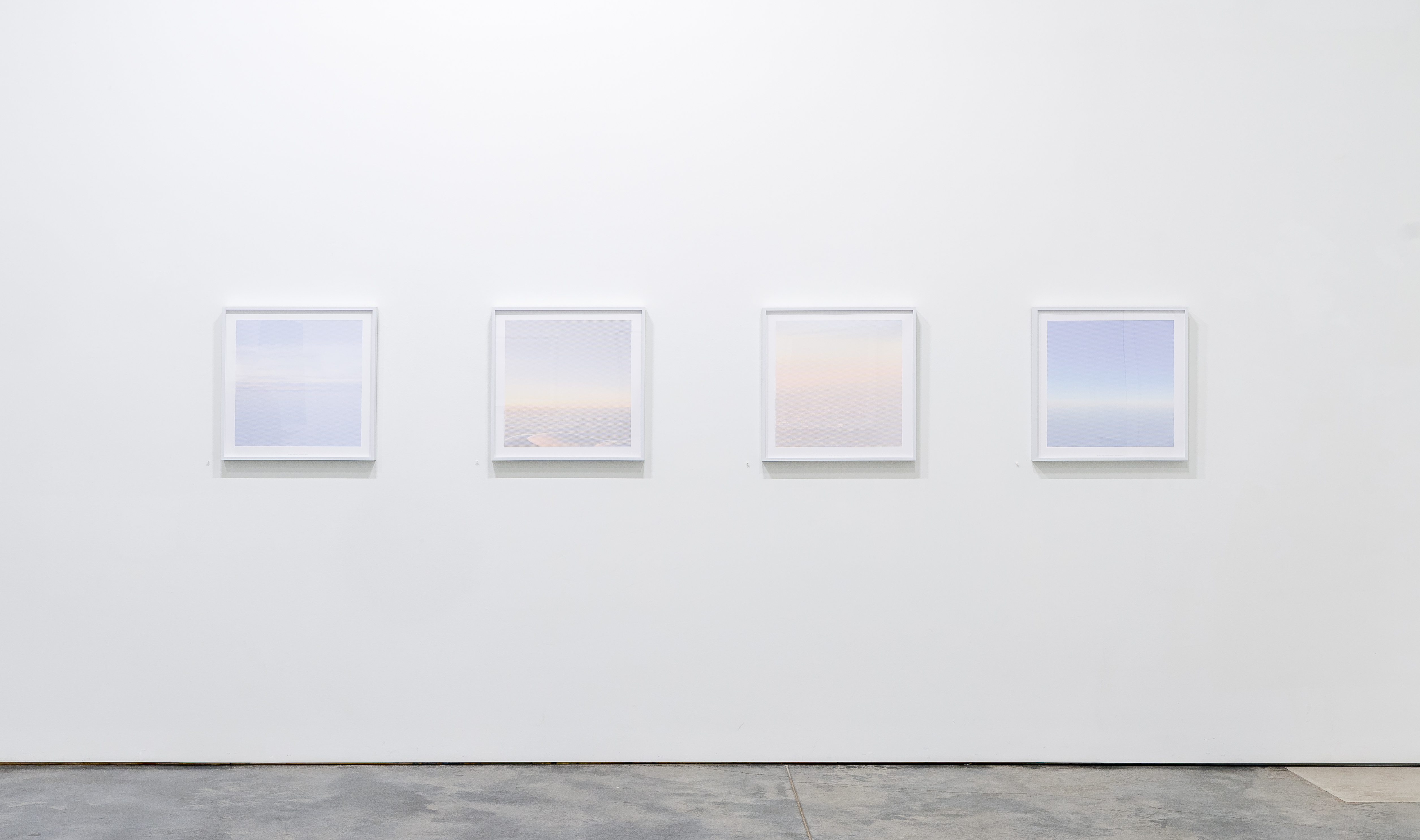 Four framed photographs arranged horizontally on a white gallery wall, each depicting serene seascapes with horizons dividing sky and water in soft gradations of blue, pink, and cream tones rendered in a dotted pointillist style.