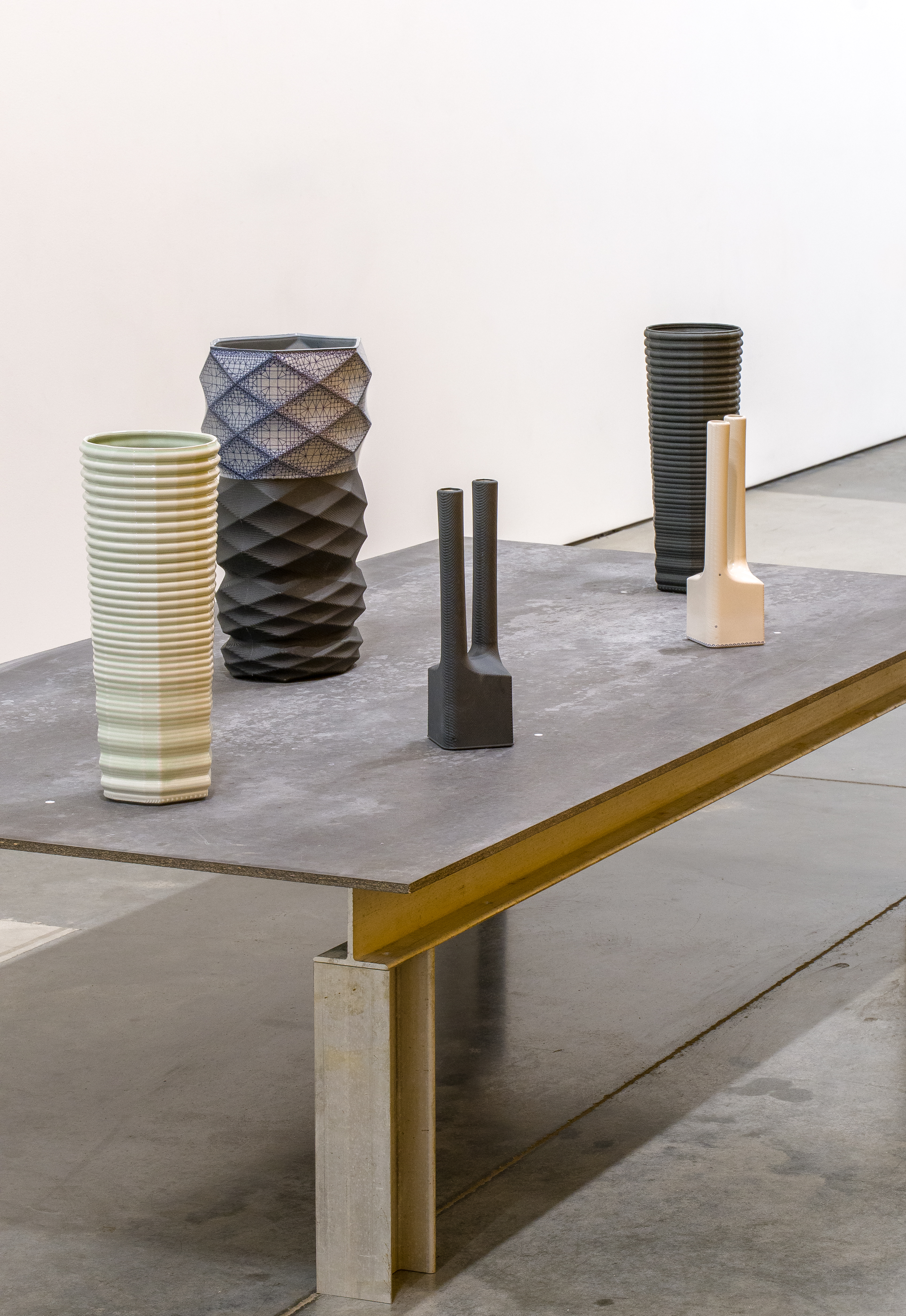 A concrete table with brass edge support displays six ceramic vessels of varying heights and textures arranged in a sparse composition. Works include a pale green ribbed cylinder, a dark patterned vessel with geometric quilting, a textured black form, two small dark cylindrical pieces, a tall ribbed dark vessel, and a cream-colored cubic base with extending form. The minimal arrangement emphasizes the individual materiality and surface treatment of each sculptural object against a neutral gallery backdrop.