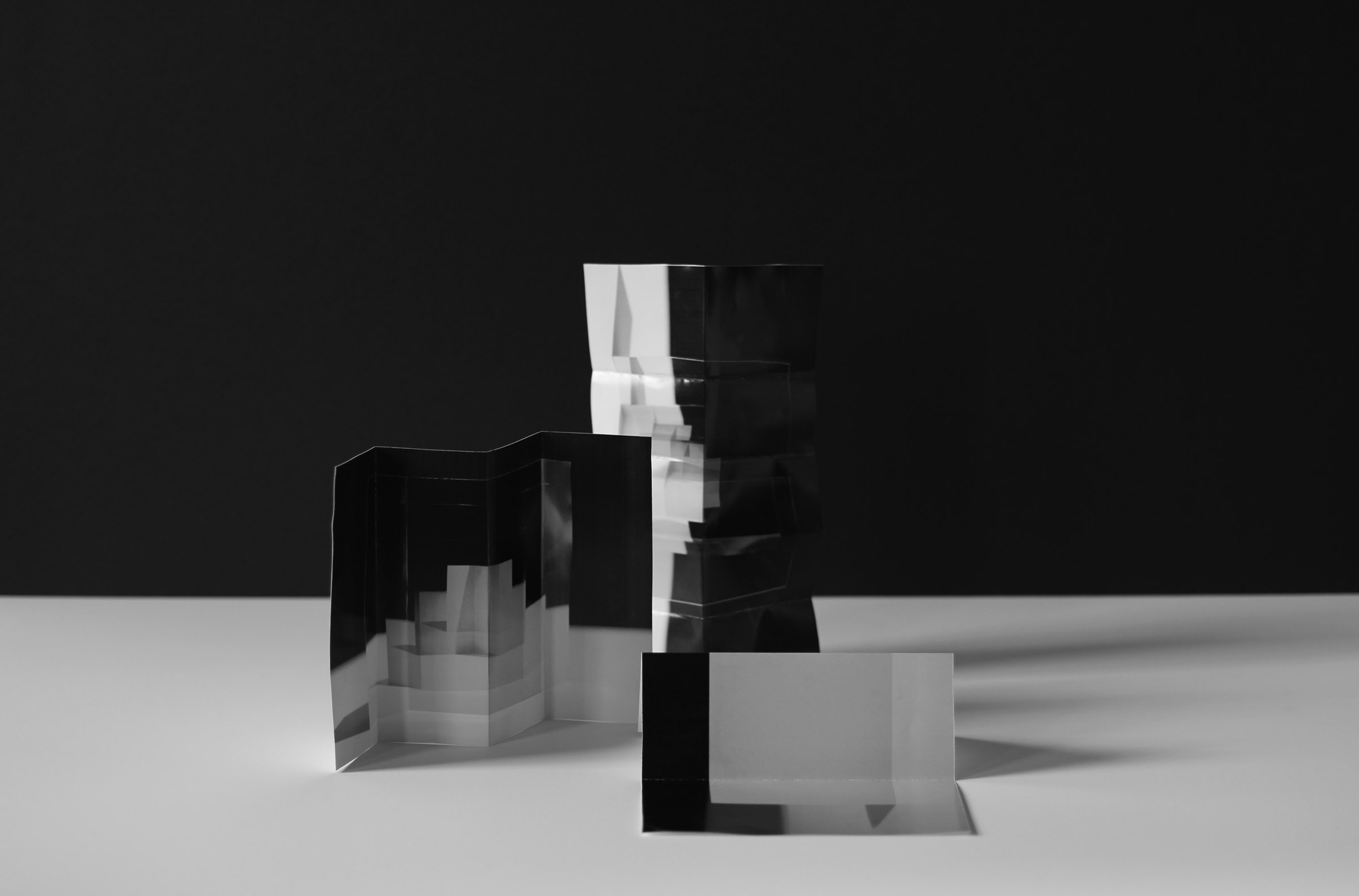 Three-dimensional paper or card structures with black and white surfaces arranged on a light gray floor against a black wall, creating geometric forms with layered planes and sharp angles that cast distinct shadows.