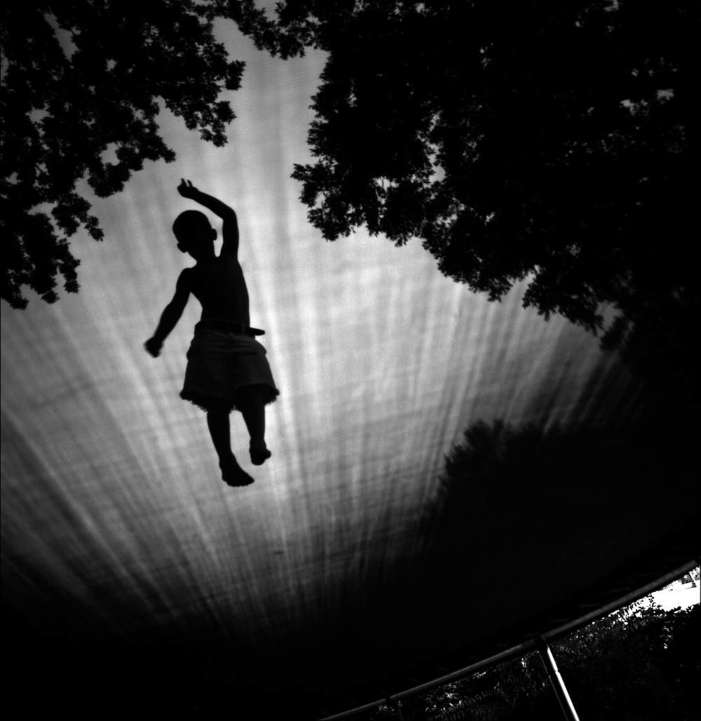 Silhouette of a child mid-jump on a trampoline, captured from below against a bright sky. Dark tree foliage frames the top and sides of the composition, creating a striking contrast with the luminous center in this black and white photograph.