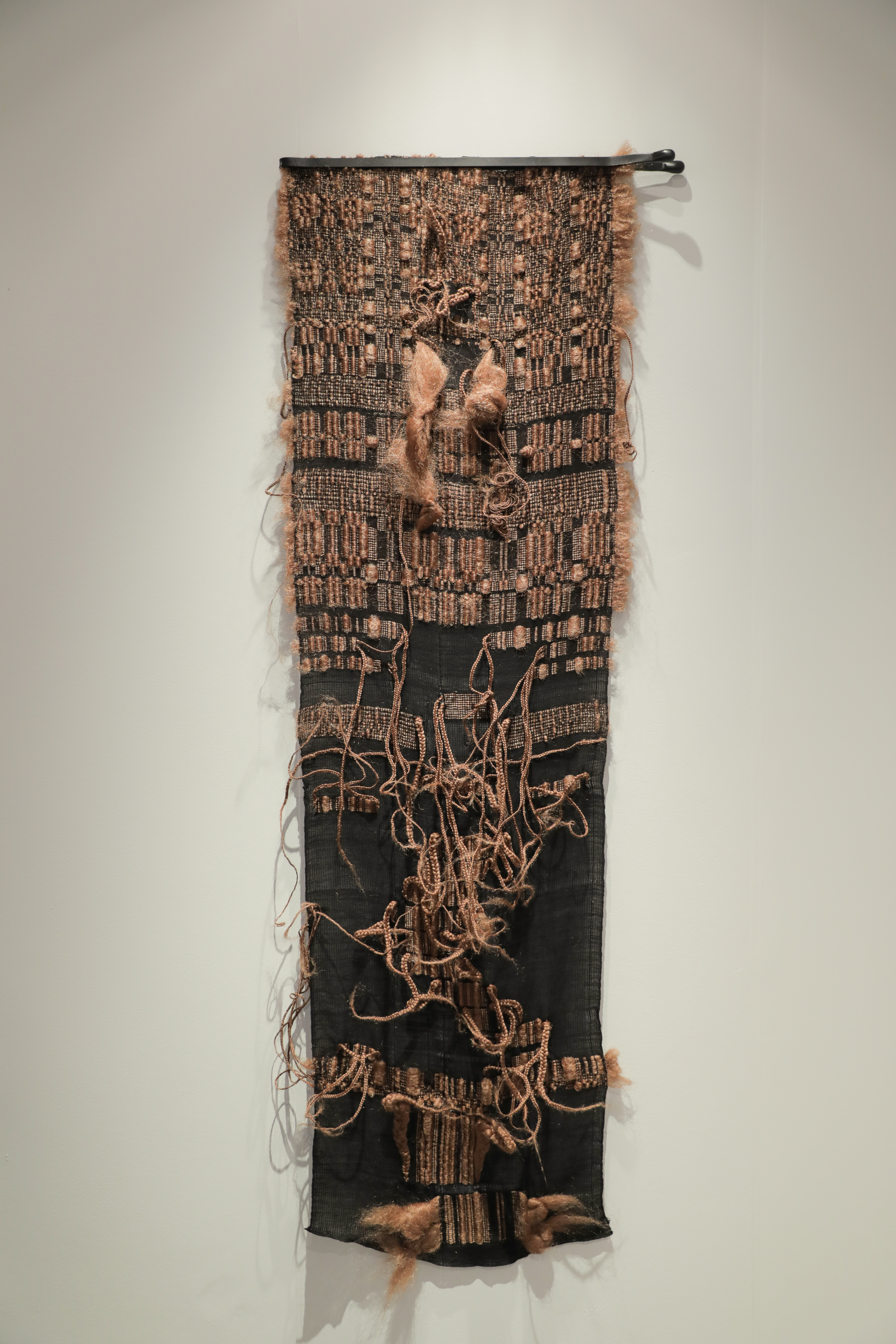 A vertical textile wall hanging combining woven cotton with synthetic hair elements. The upper portion features a tan and black woven pattern with figurative forms, while the lower section is predominantly black with looping strands of blonde hair creating organic, gestural marks across the surface.