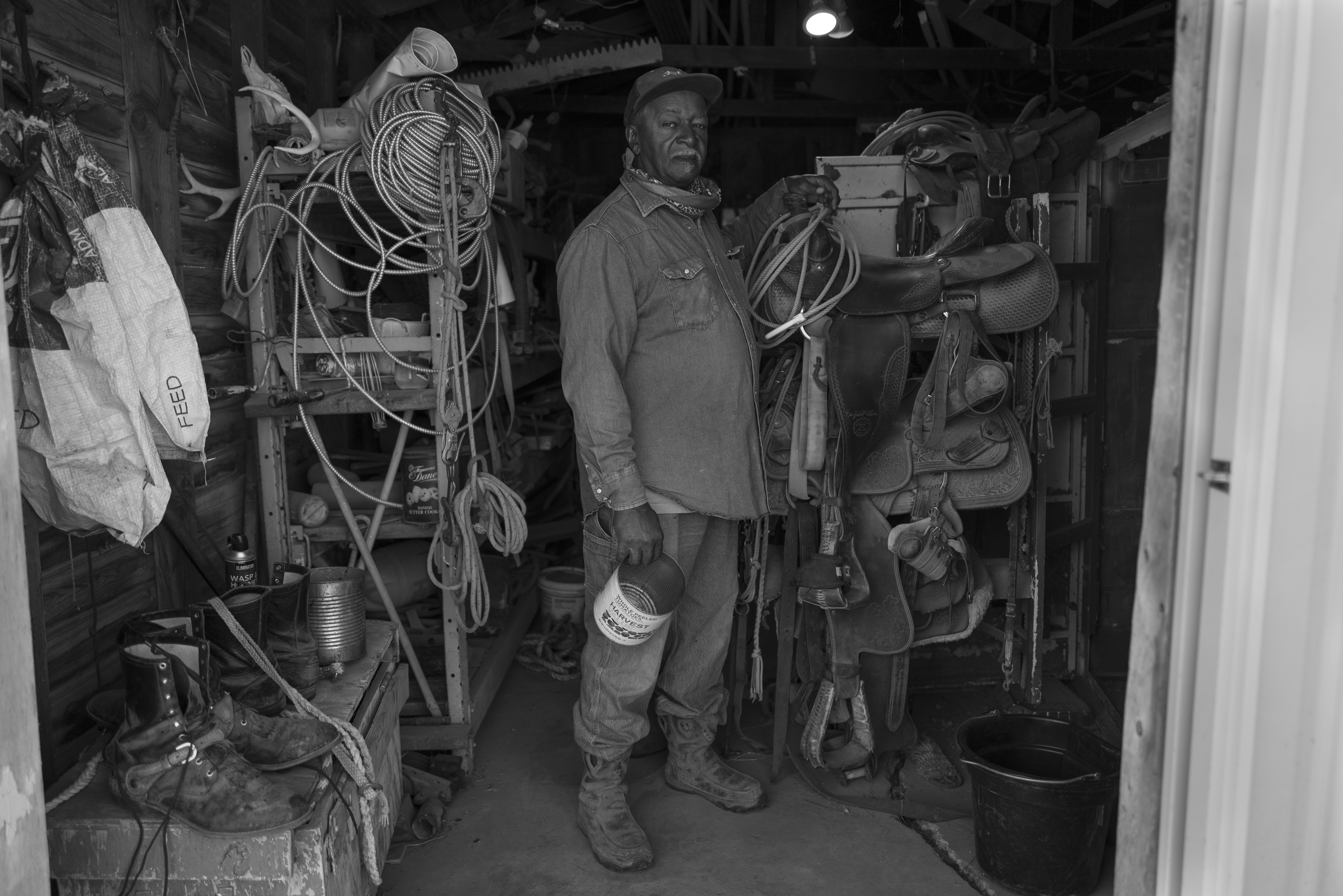 Black and white photograph depicting a man in work clothes and hat standing in a cluttered storage or workshop space surrounded by coiled ropes, saddles, leather equipment, and various ranch tools hanging on walls and mounted on stands.