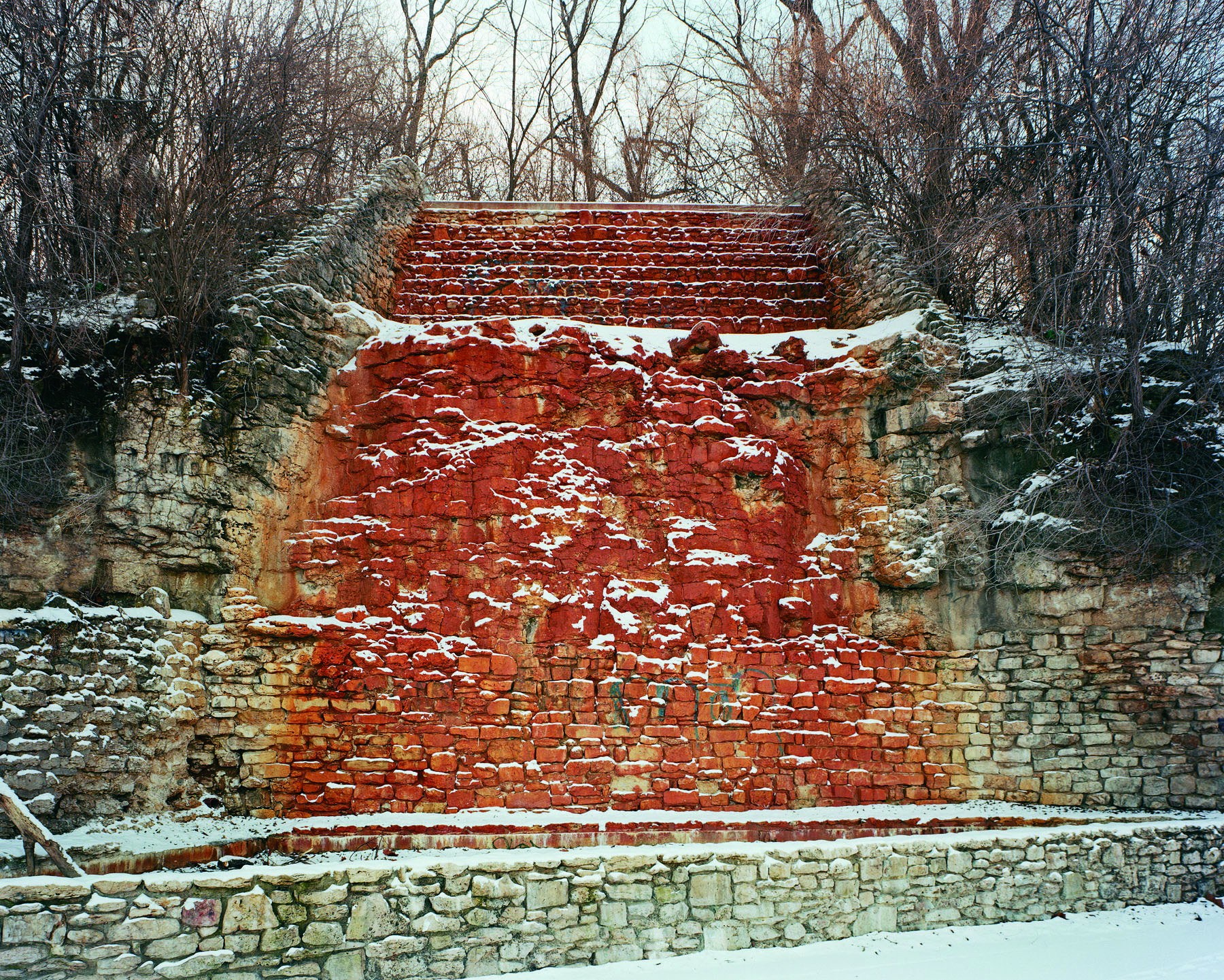 A dramatic red brick structure with stepped architectural forms rises from a snowy landscape, flanked by bare winter trees and stone walls. Patches of snow cling to the vivid red and orange brickwork, creating stark contrast against the muted forest backdrop.