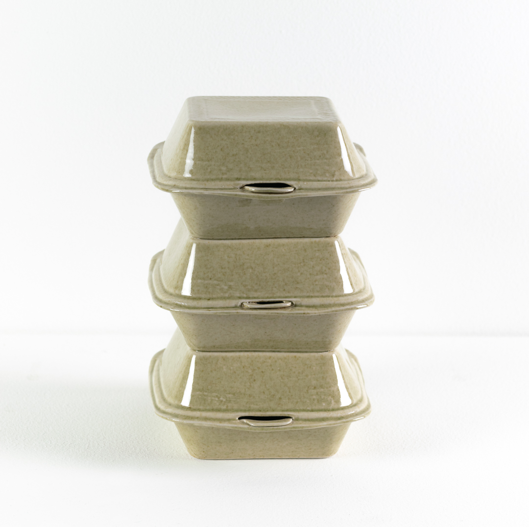 Three stacked porcelain takeout containers with hinged lids in pale celadon green, arranged vertically to form a tower-like sculptural form against a white background.