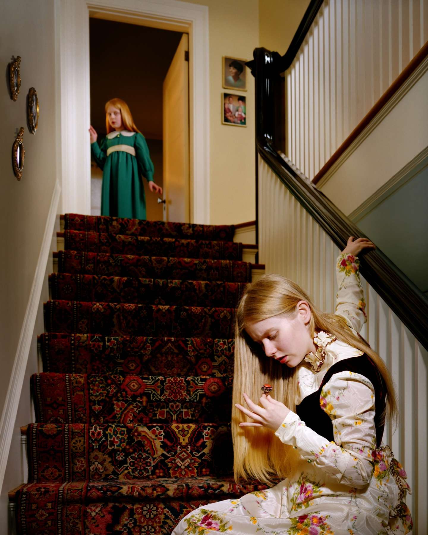 A young girl in a floral dress sits on a carpeted staircase, examining a ring on her hand. Behind her on the stairs stands another girl in a green dress, while a dark silhouette looms against the upper landing. The scene is rendered in rich, saturated colors with warm cream and yellow walls, ornate decorative details, and patterned carpet in deep reds and golds.